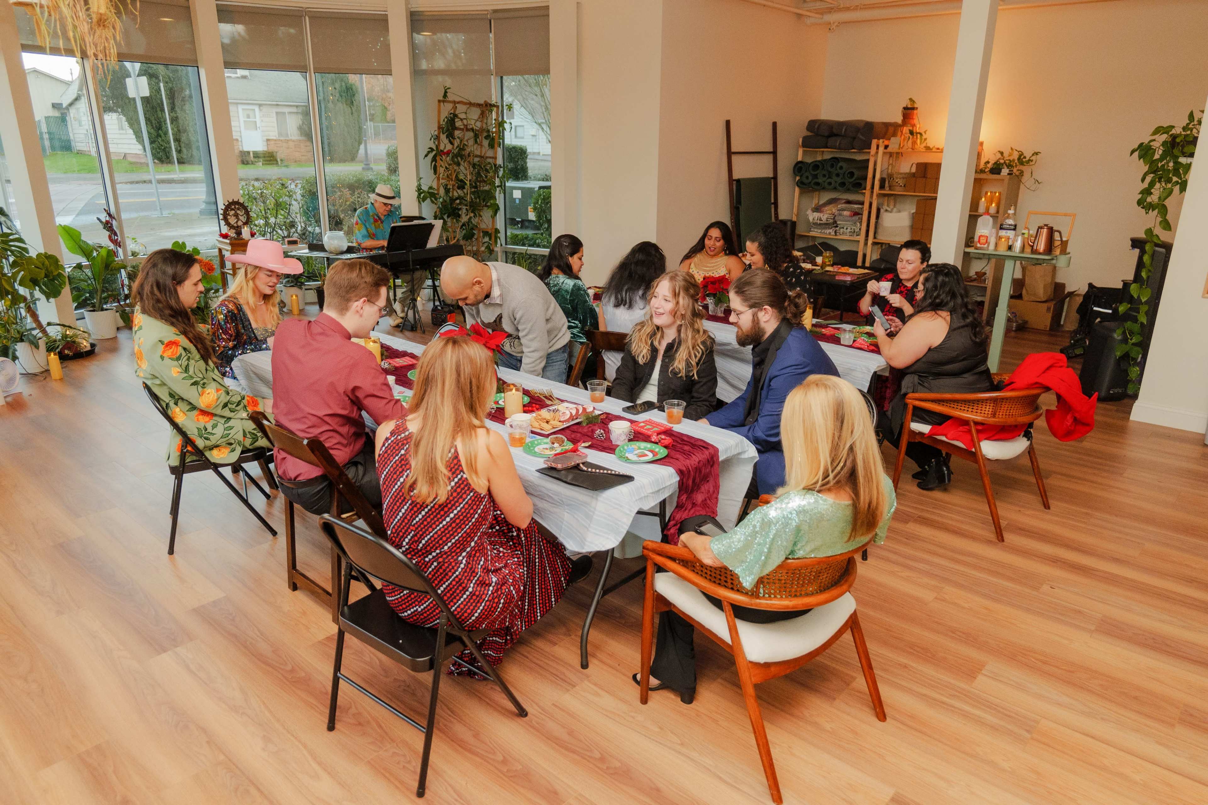 A gathering of people sits at a long table adorned with colorful plates and decorations in a well-lit room filled with plants and cozy furniture.