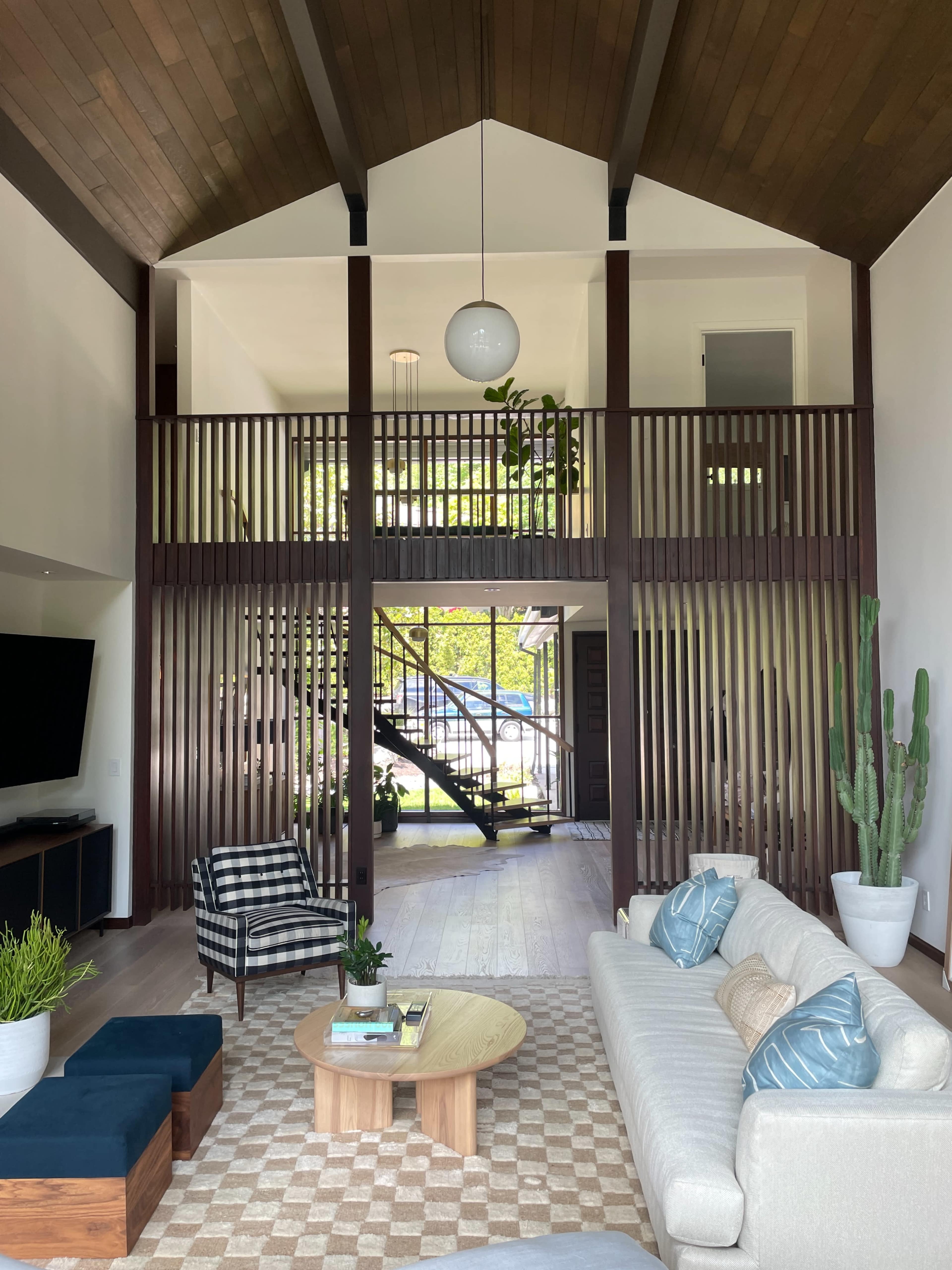 A modern living room with a high ceiling, featuring a sofa, a circular coffee table, a patterned rug, and a staircase visible in the background, separated by slatted wooden partitions.
