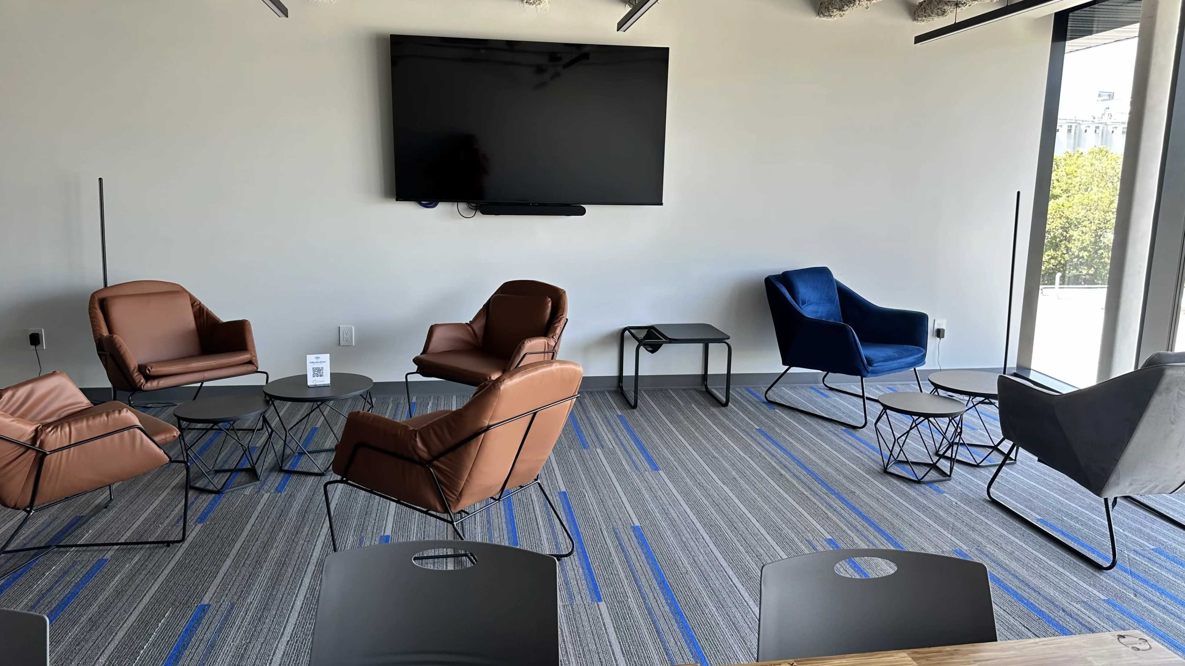 The image shows a modern meeting space with various chairs and tables arranged around a large wall-mounted television.