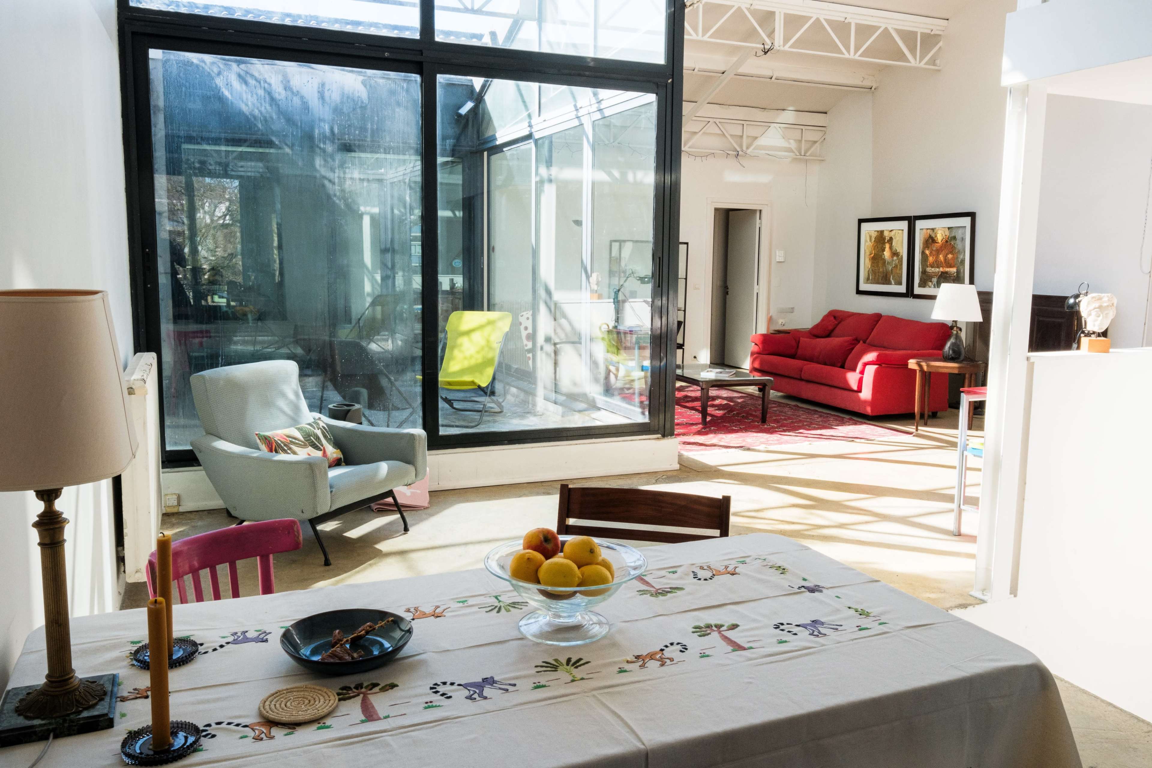 A dining table with a floral tablecloth is set in the foreground, while a bright living area with a red sofa and large windows is visible in the background.
