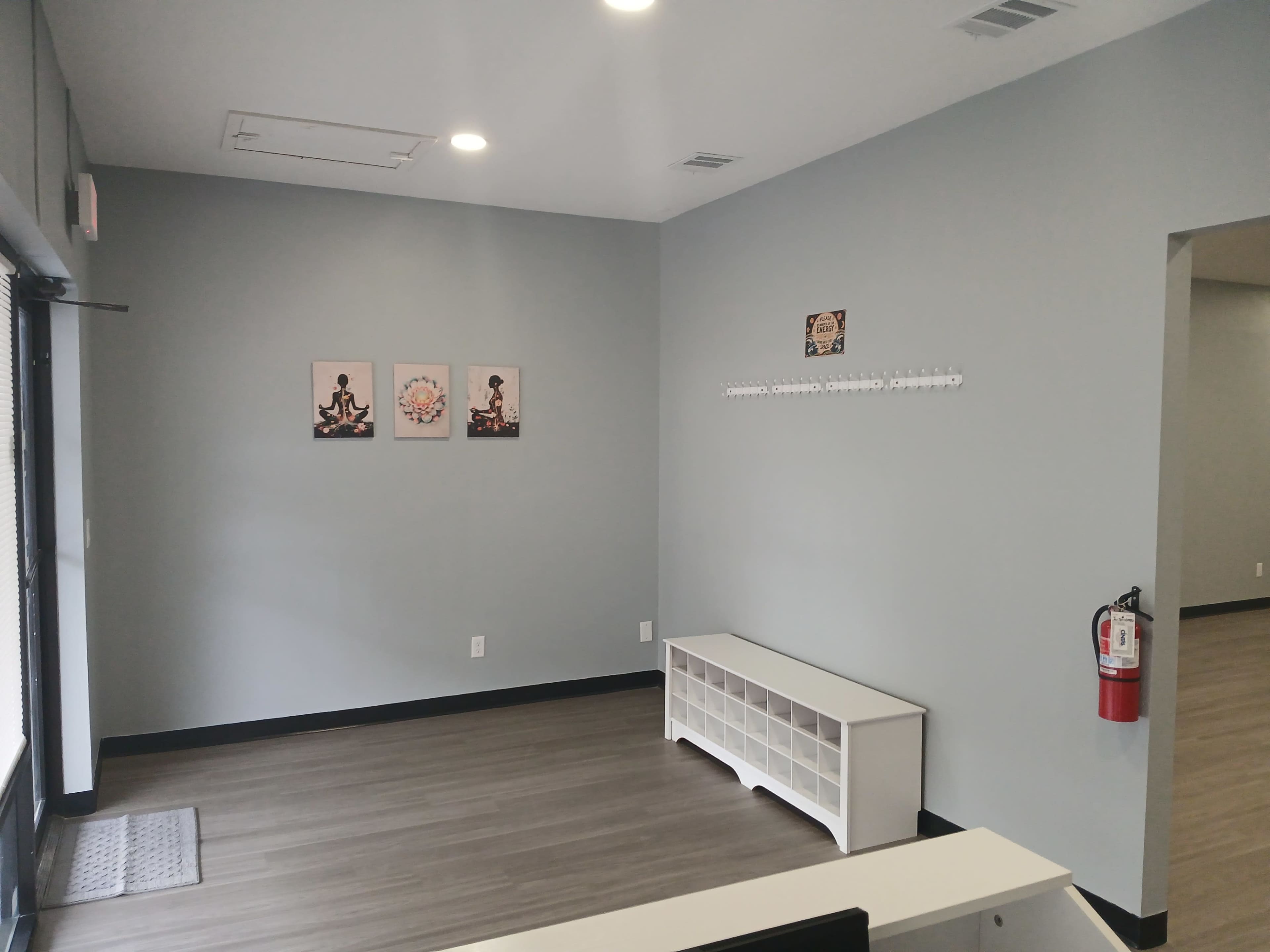 The image shows a minimalistic interior of a commercial space with light gray walls, a white storage bench, and three framed artworks on the wall.