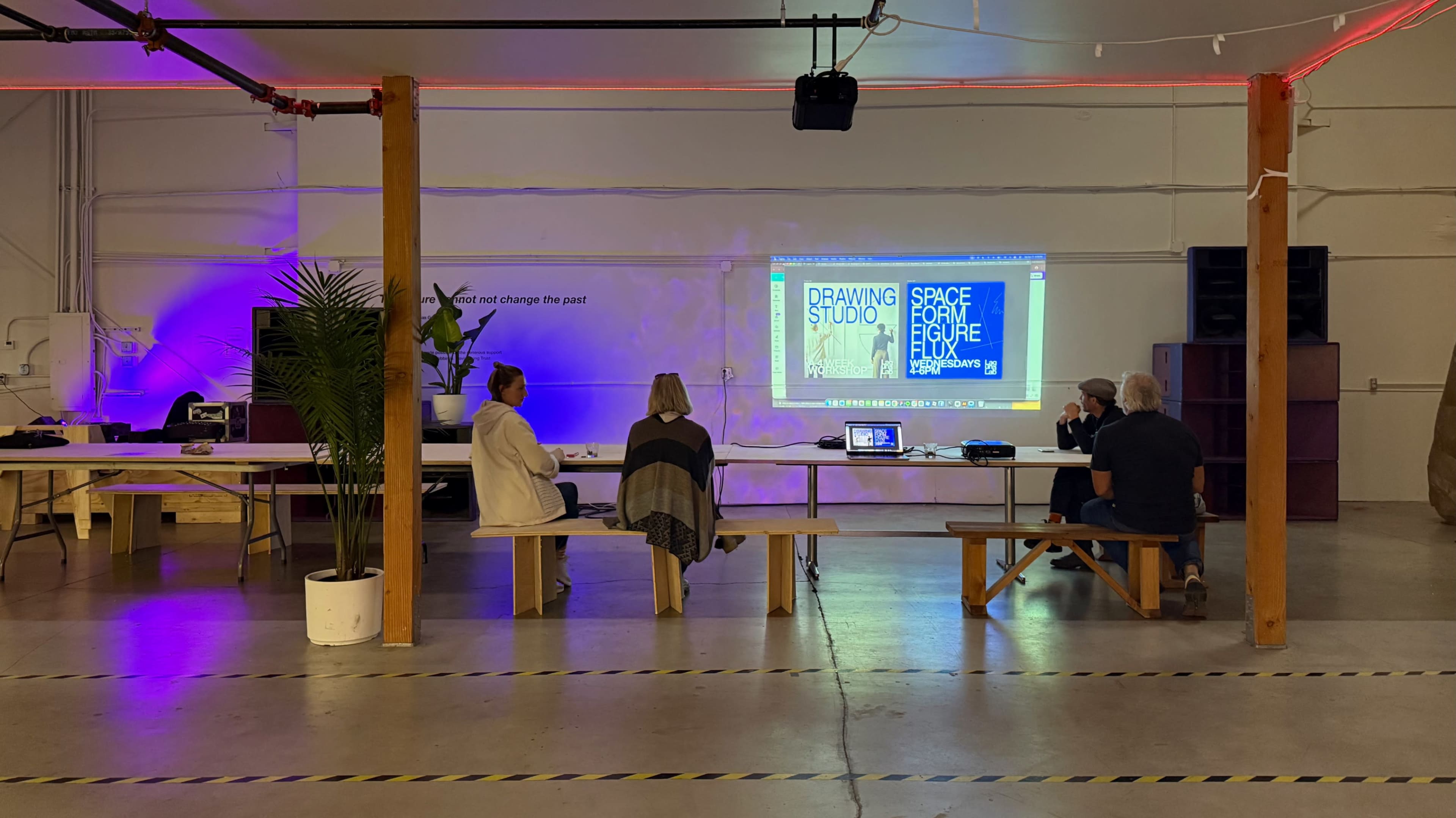 A group of five people sits on benches in a spacious room with a large screen displaying text, while a plant and various tables set up for an event are visible.