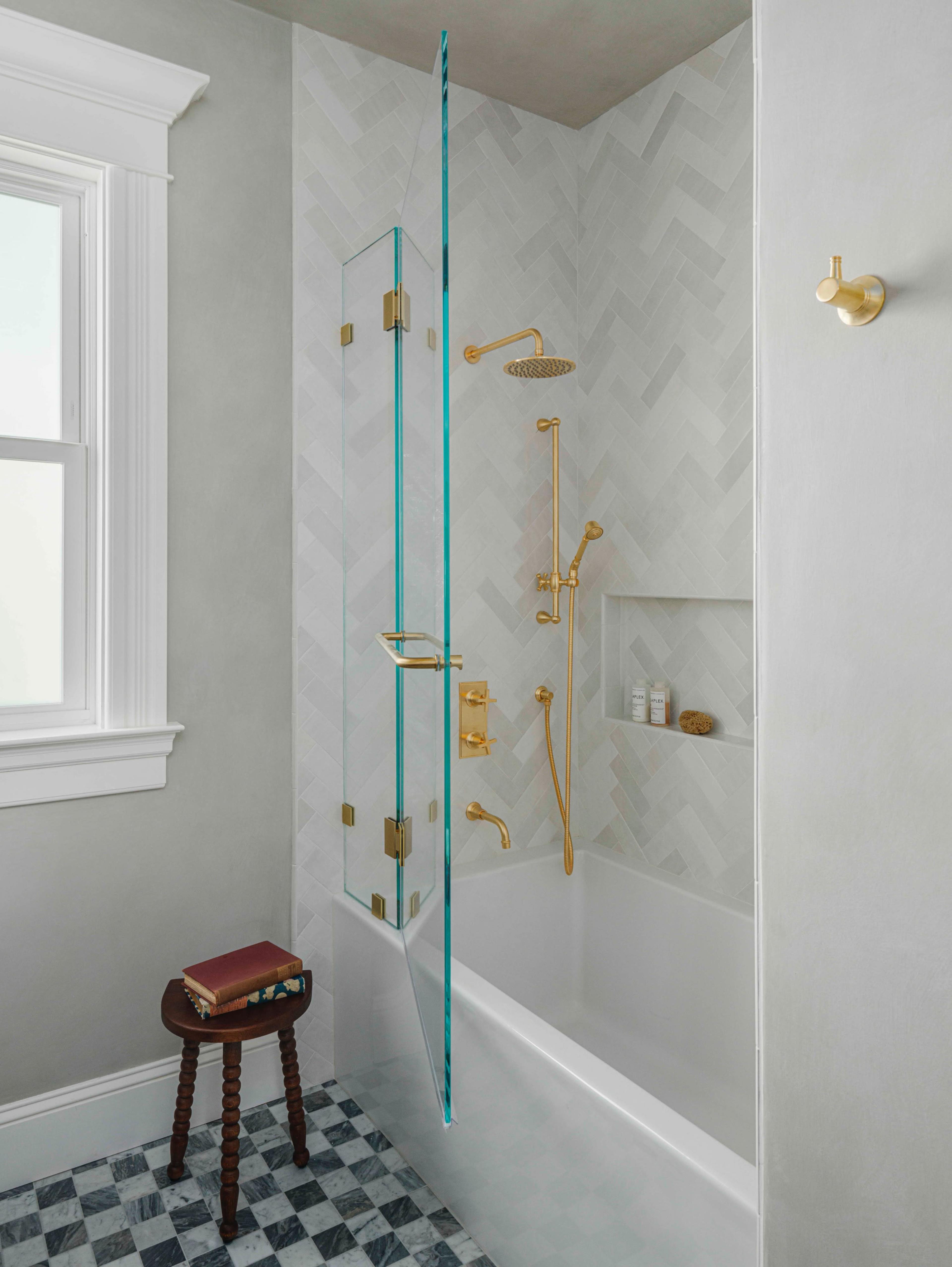 The image shows a modern shower with a glass enclosure, gold fittings, and a herringbone tile pattern on the walls, located in a bathroom with a window and a small stool beside a bathtub.