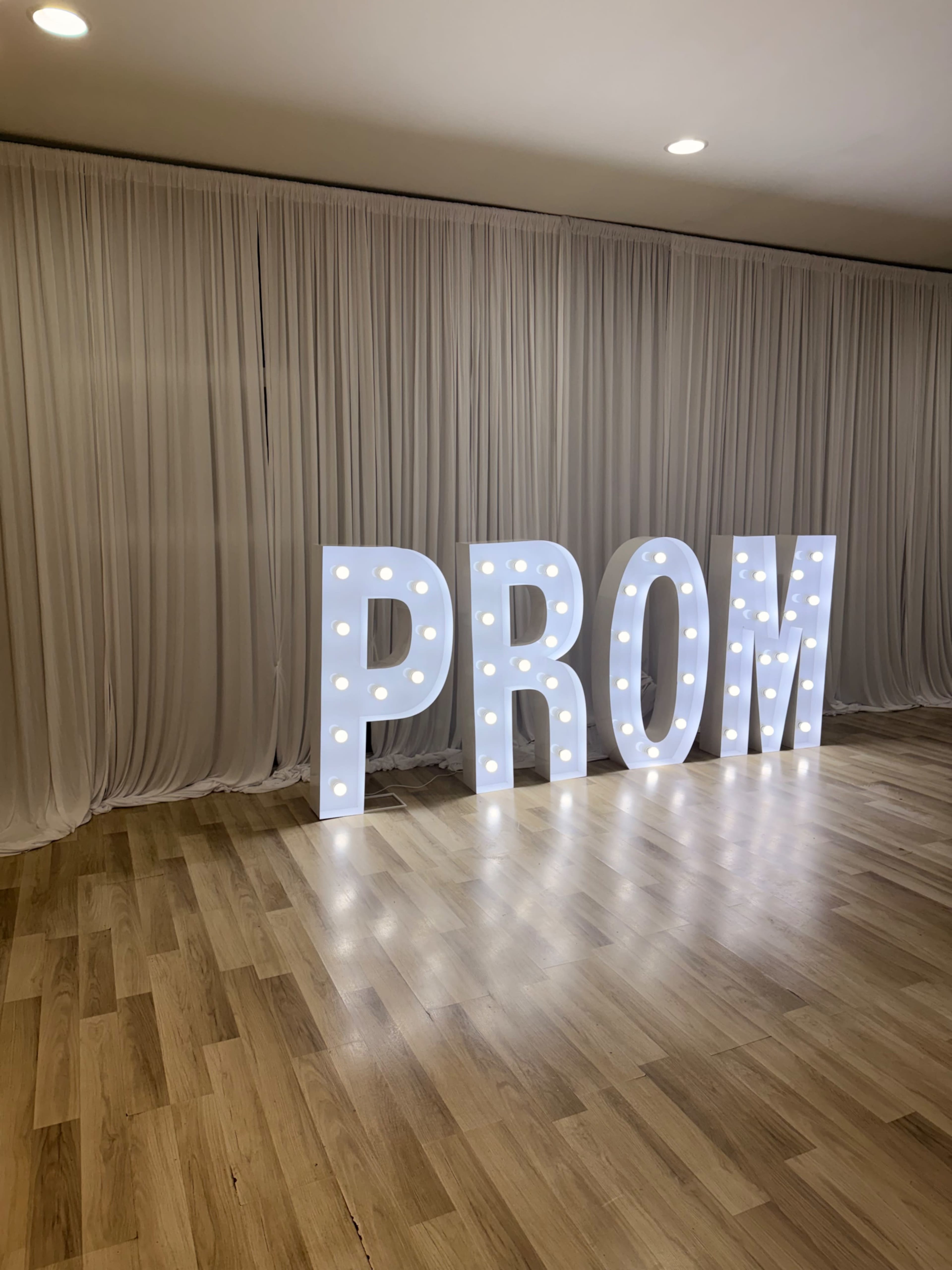 The image shows large, illuminated letters spelling "PROM" in front of a backdrop of white draped fabric.