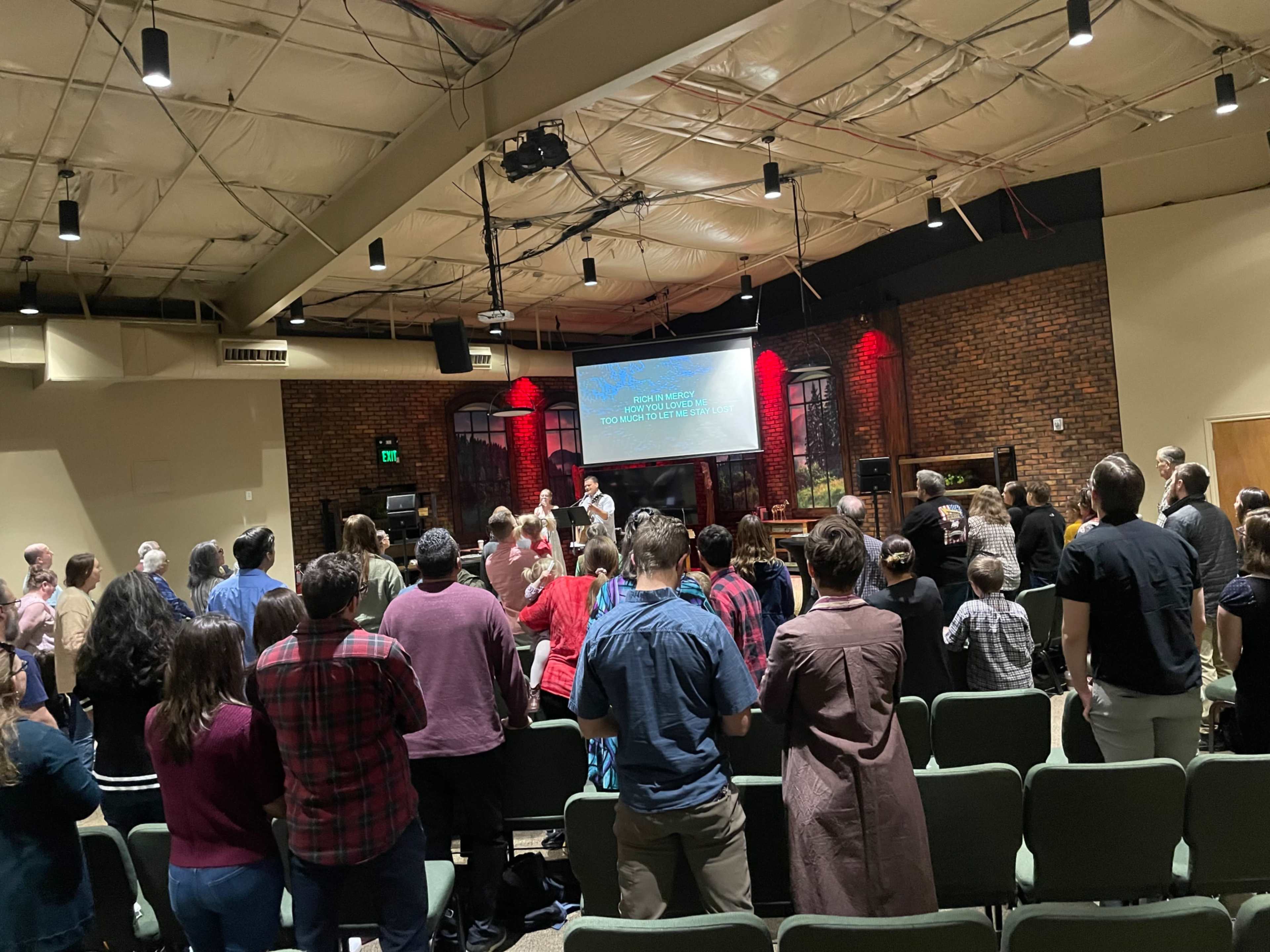 A congregation stands in a church, facing a large screen displaying text, while the service is in progress.