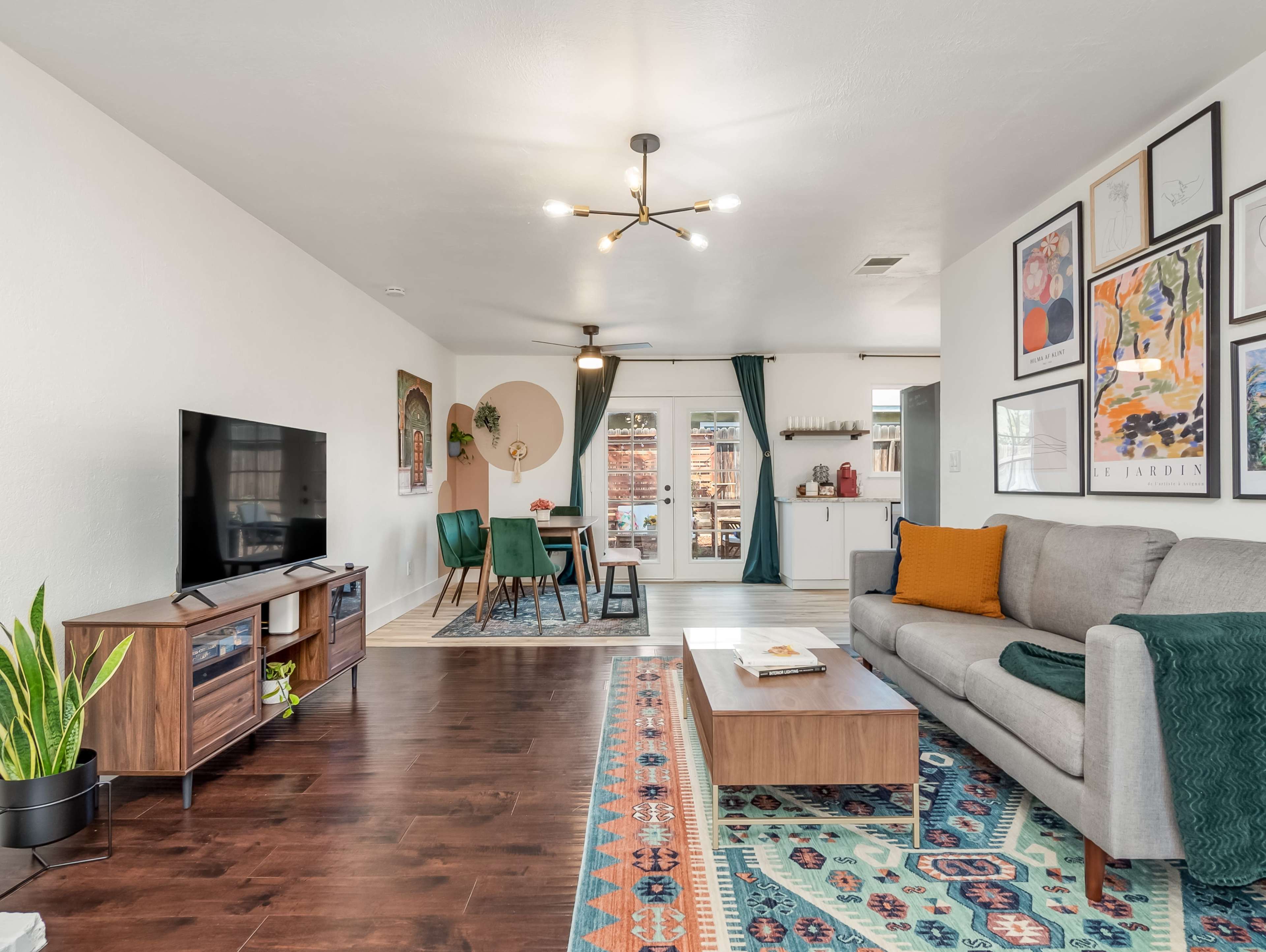 The living room features a gray sofa, a wooden coffee table, a TV stand with a television, and a dining area in the background, all accented by a colorful area rug and large windows.