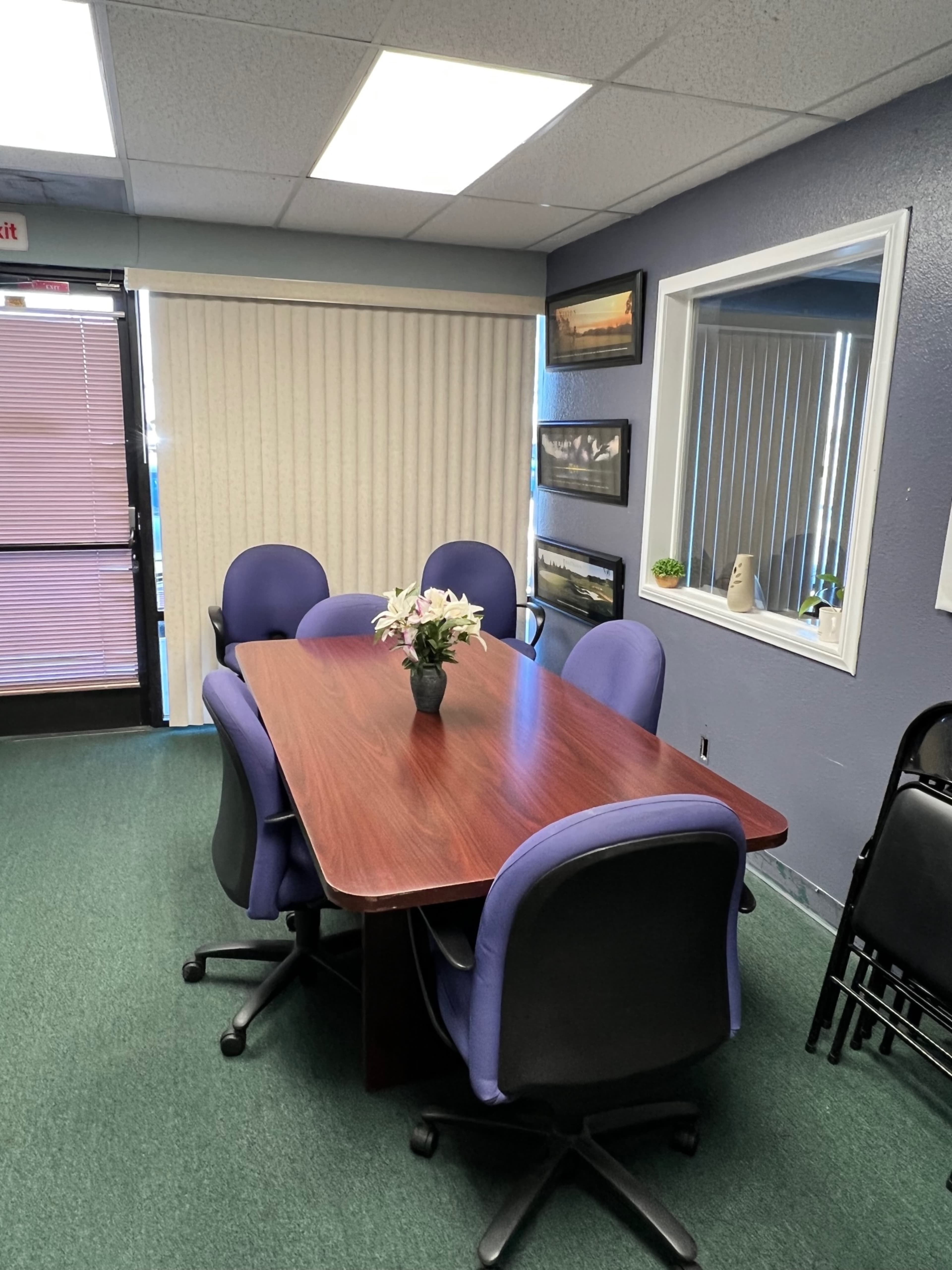 A conference room with a wooden table surrounded by purple chairs, featuring a flower centerpiece and a window with blinds.