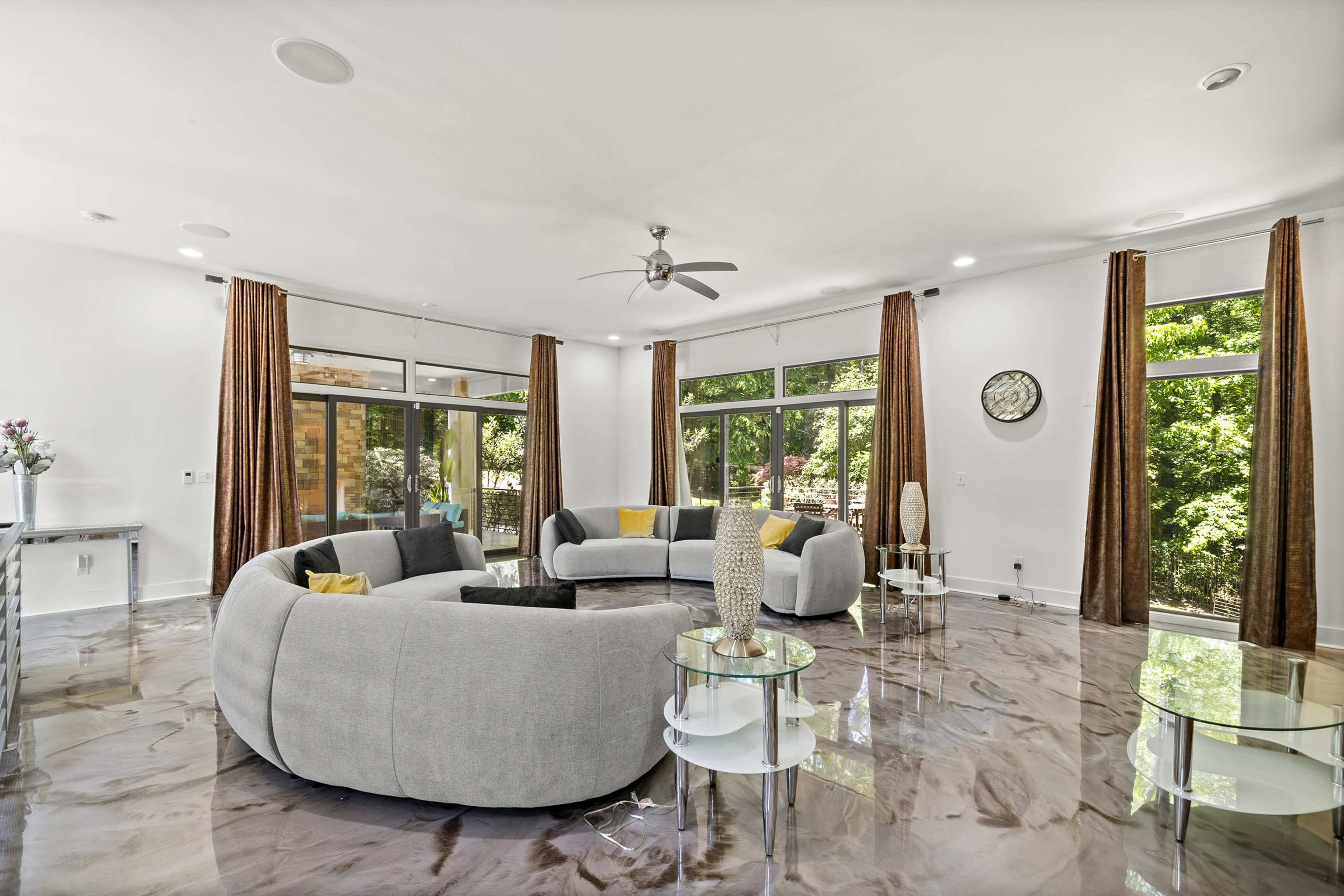 The image displays a modern living room featuring grey circular sofas, glass coffee tables, and large windows allowing natural light, with greenery visible outside.