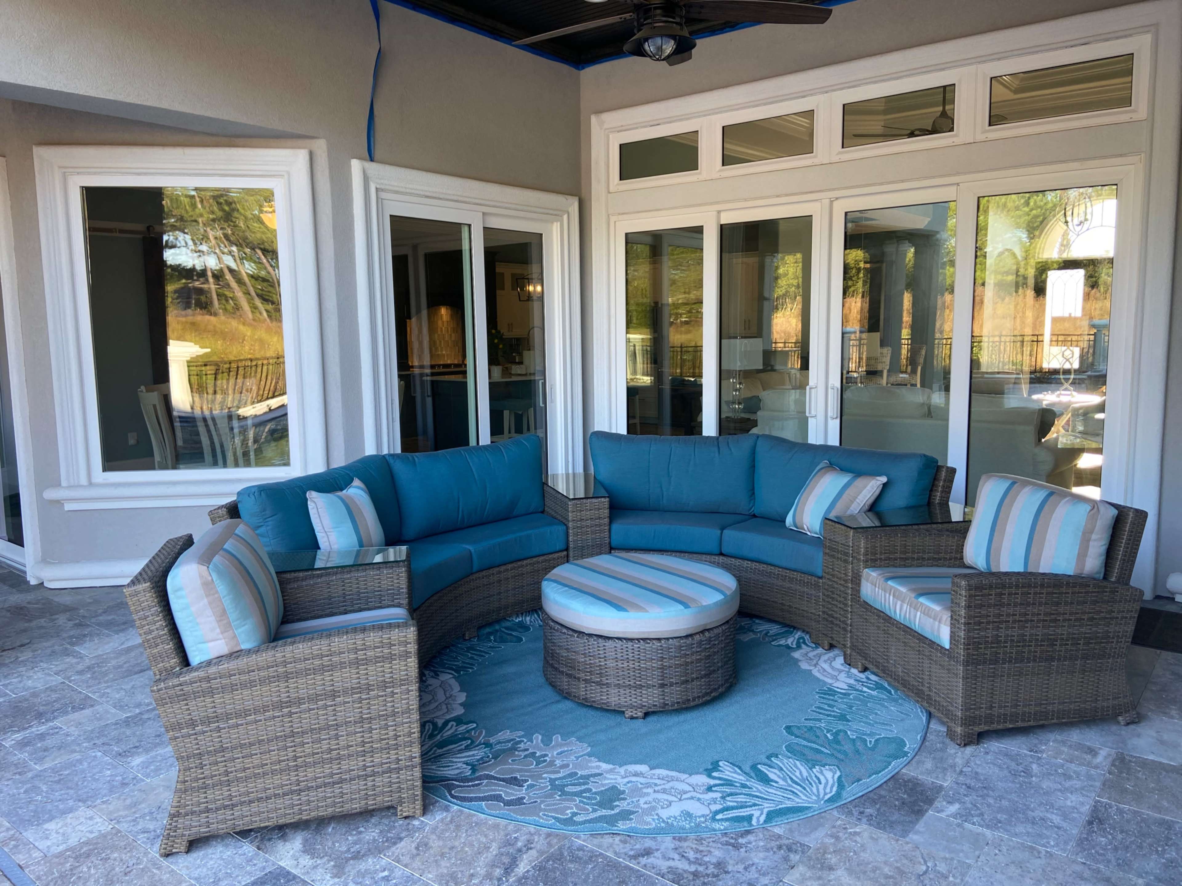 A patio area featuring a circular seating arrangement in blue cushions, a matching round coffee table, and a decorative rug on a tiled floor, enclosed by large glass doors.