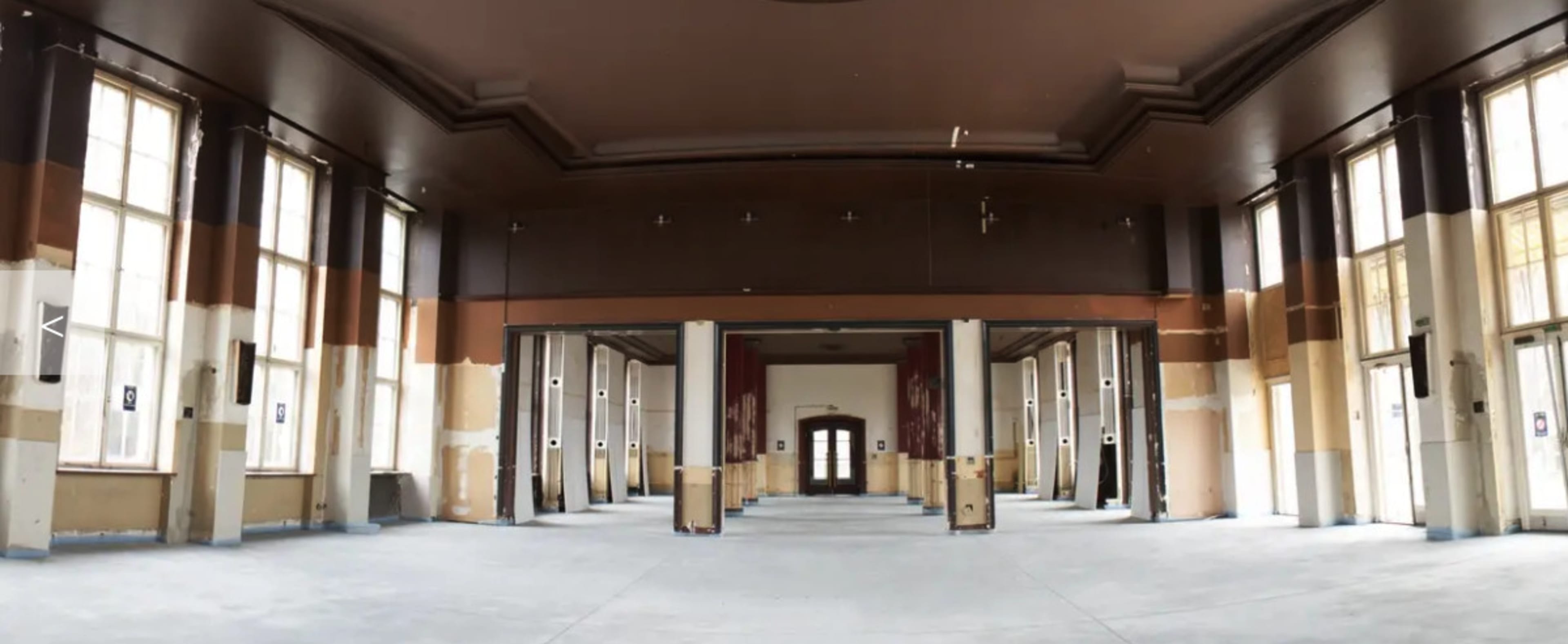 The image shows an empty, spacious interior of a building with large windows and exposed walls under renovation.