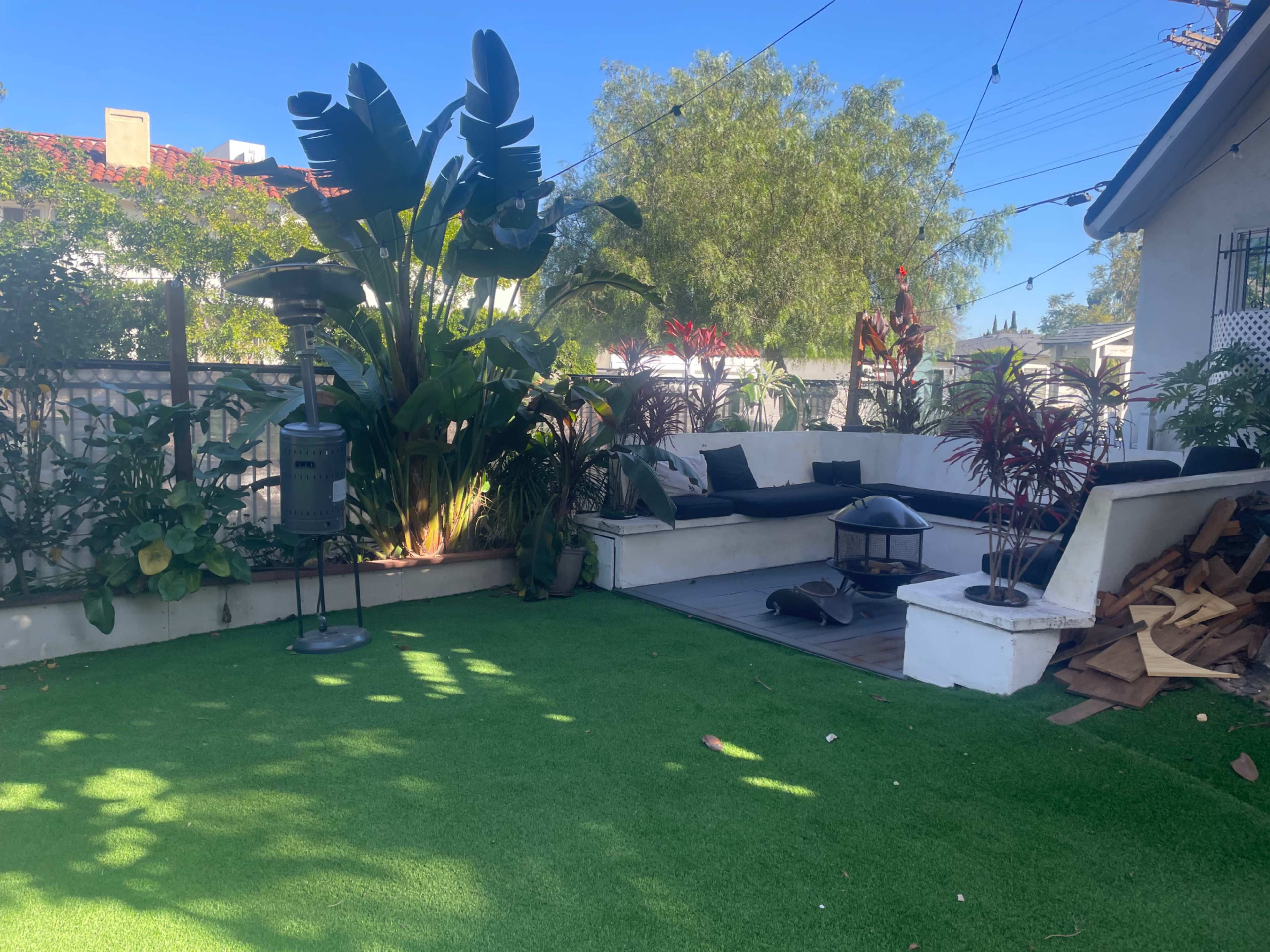 The image shows an outdoor patio area with a green artificial lawn, surrounded by various plants, a seating arrangement, and a fire pit.