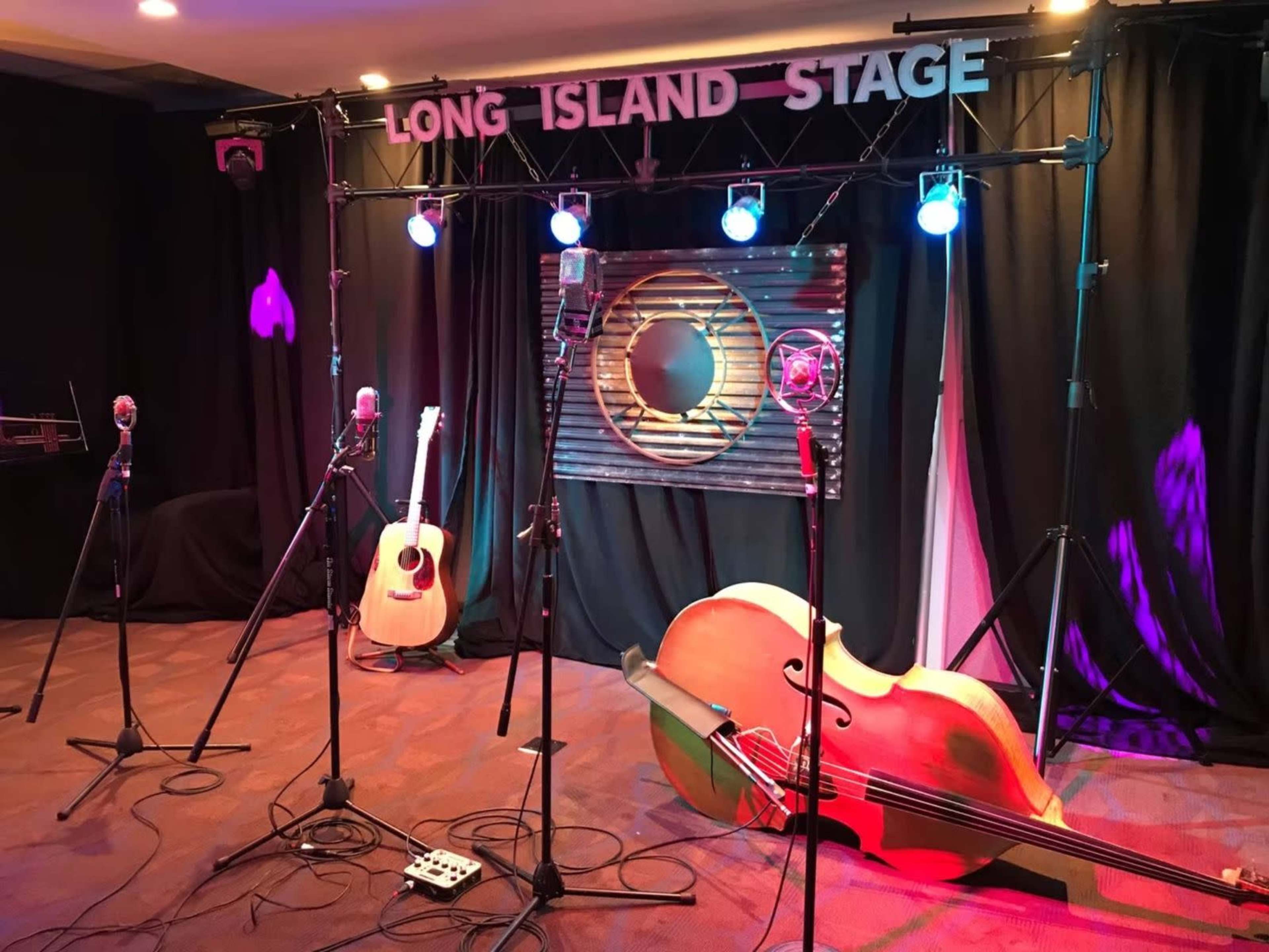 The image shows a stage setup with microphones, an acoustic guitar, and a double bass, framed by a sign that reads "LONG ISLAND STAGE" and illuminated by colorful stage lights.