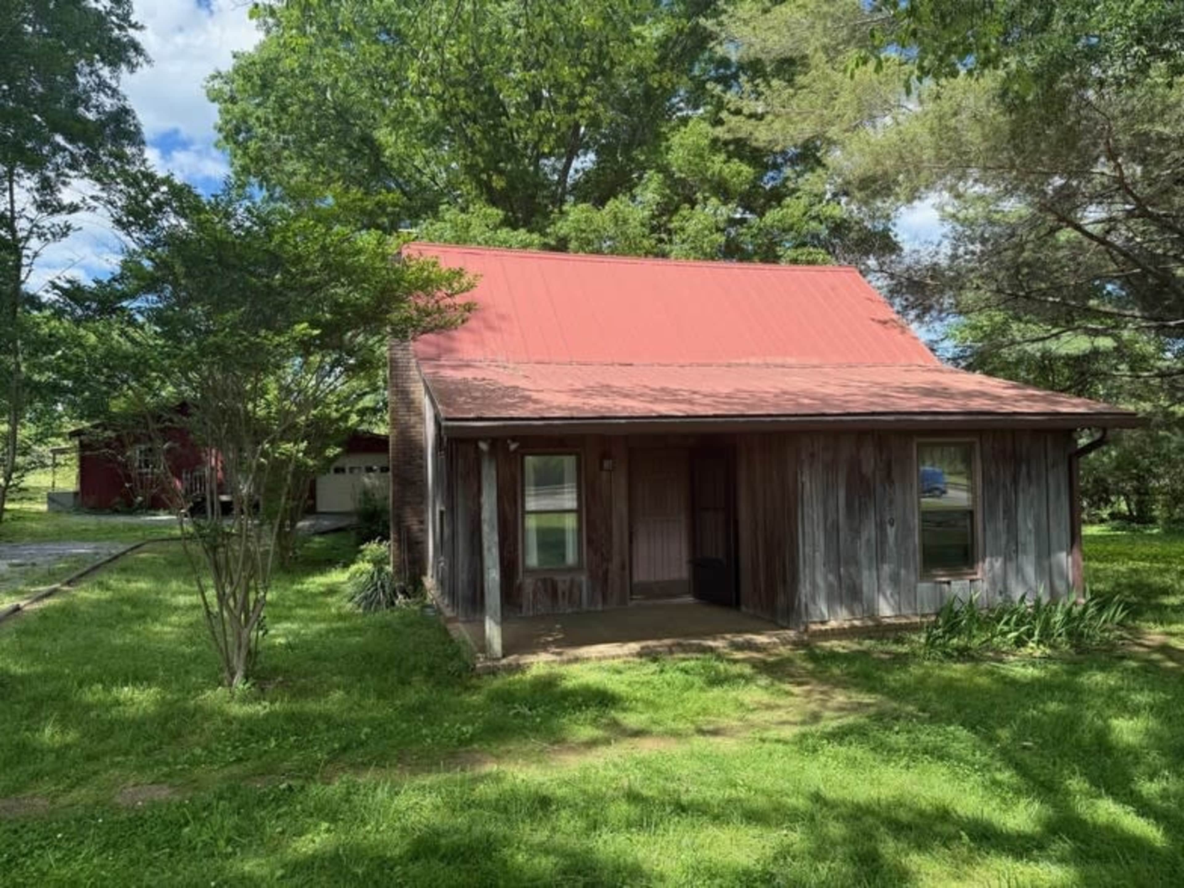 The image shows a rustic wooden cabin with a red metal roof set in a grassy area surrounded by trees.