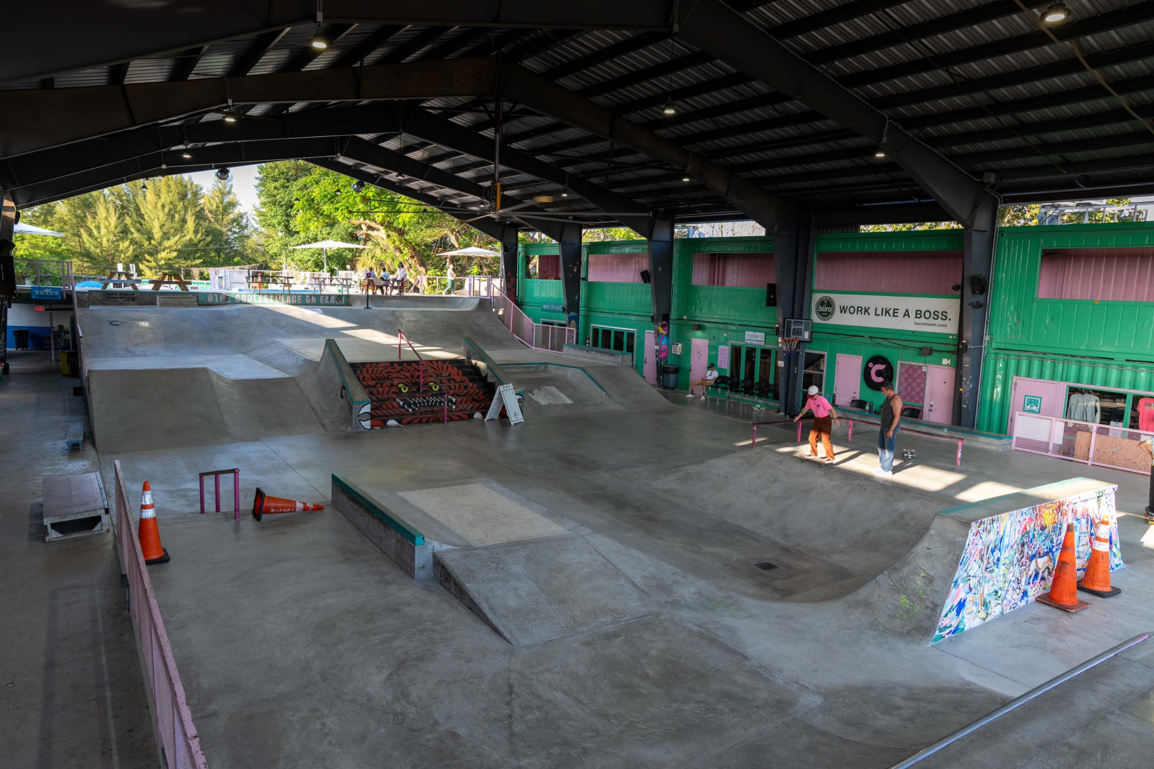 The image depicts a large indoor skatepark featuring multiple ramps, rails, and a concrete surface under a metal roof.