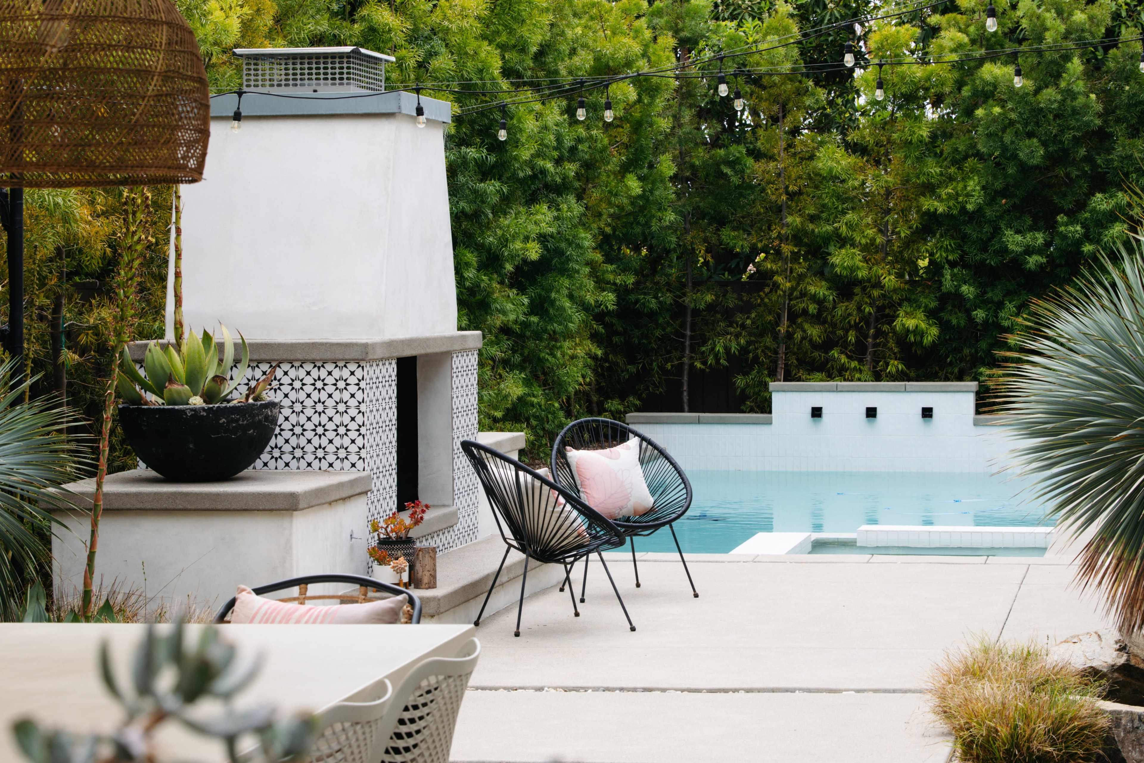 The image shows a modern outdoor space featuring a pool, a fireplace, and black wicker chairs situated near greenery.