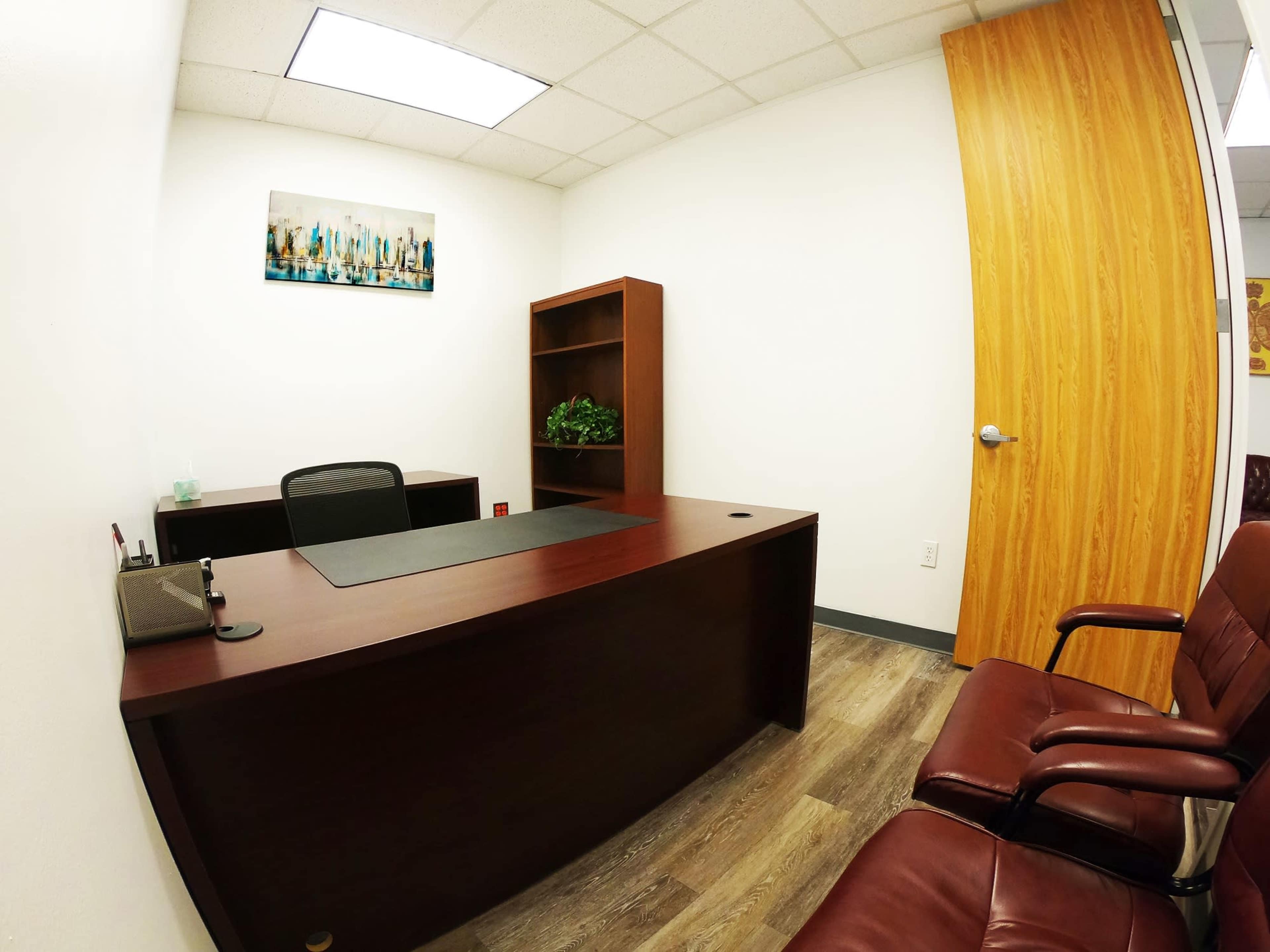 The image shows a small office with a wooden desk, a chair, a bookshelf, and a door leading outside.