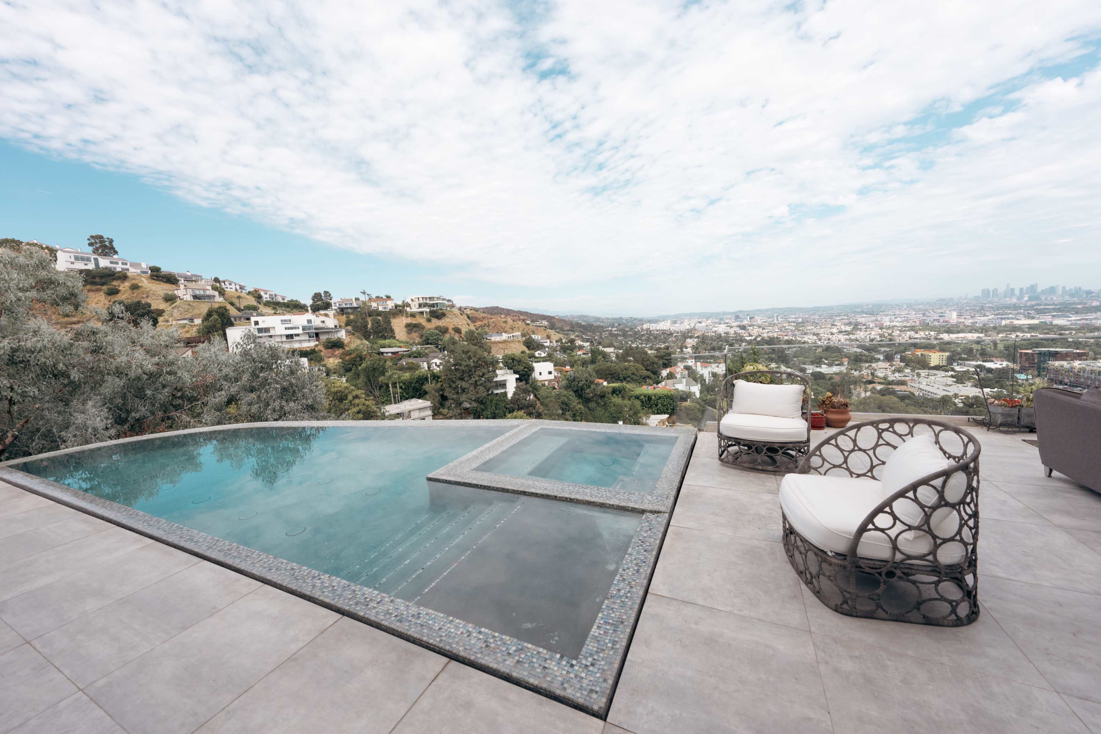 The image shows a modern pool area overlooking a cityscape with hills, featuring lounge chairs and a clear sky.