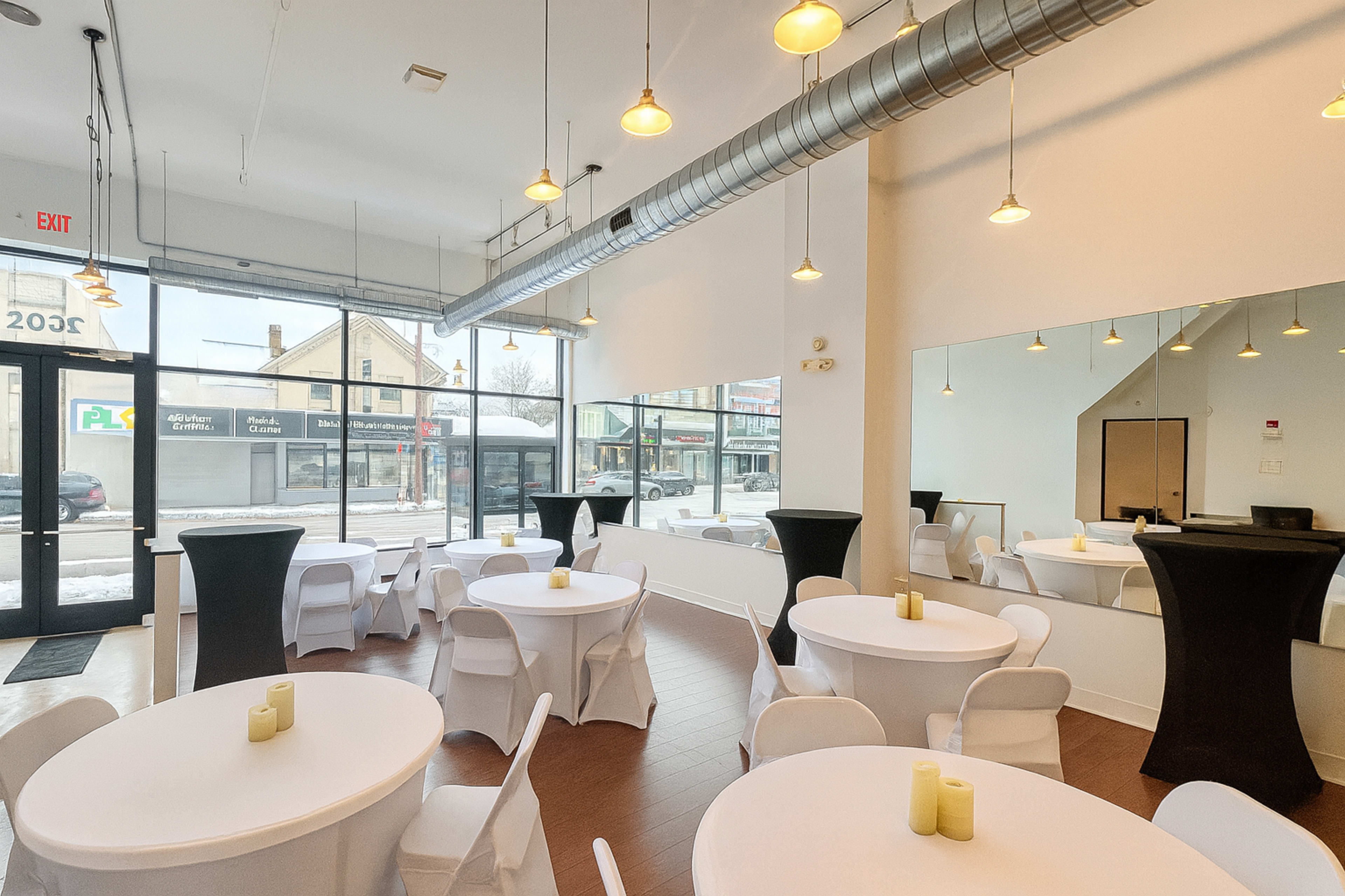 The image shows a modern event space with round tables dressed in white tablecloths and tall black tables, illuminated by pendant lights and featuring large windows overlooking a street.