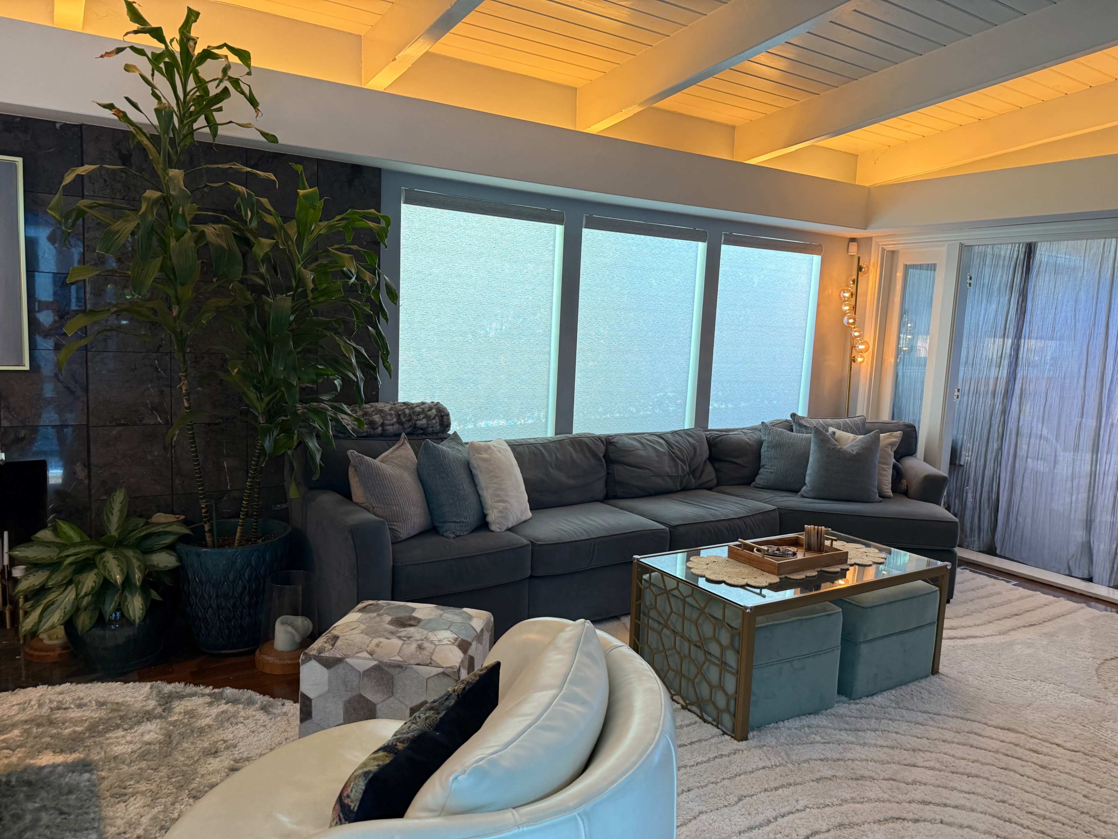 The image shows a modern living room featuring a large gray sectional sofa, a glass coffee table, and large windows with soft lighting.