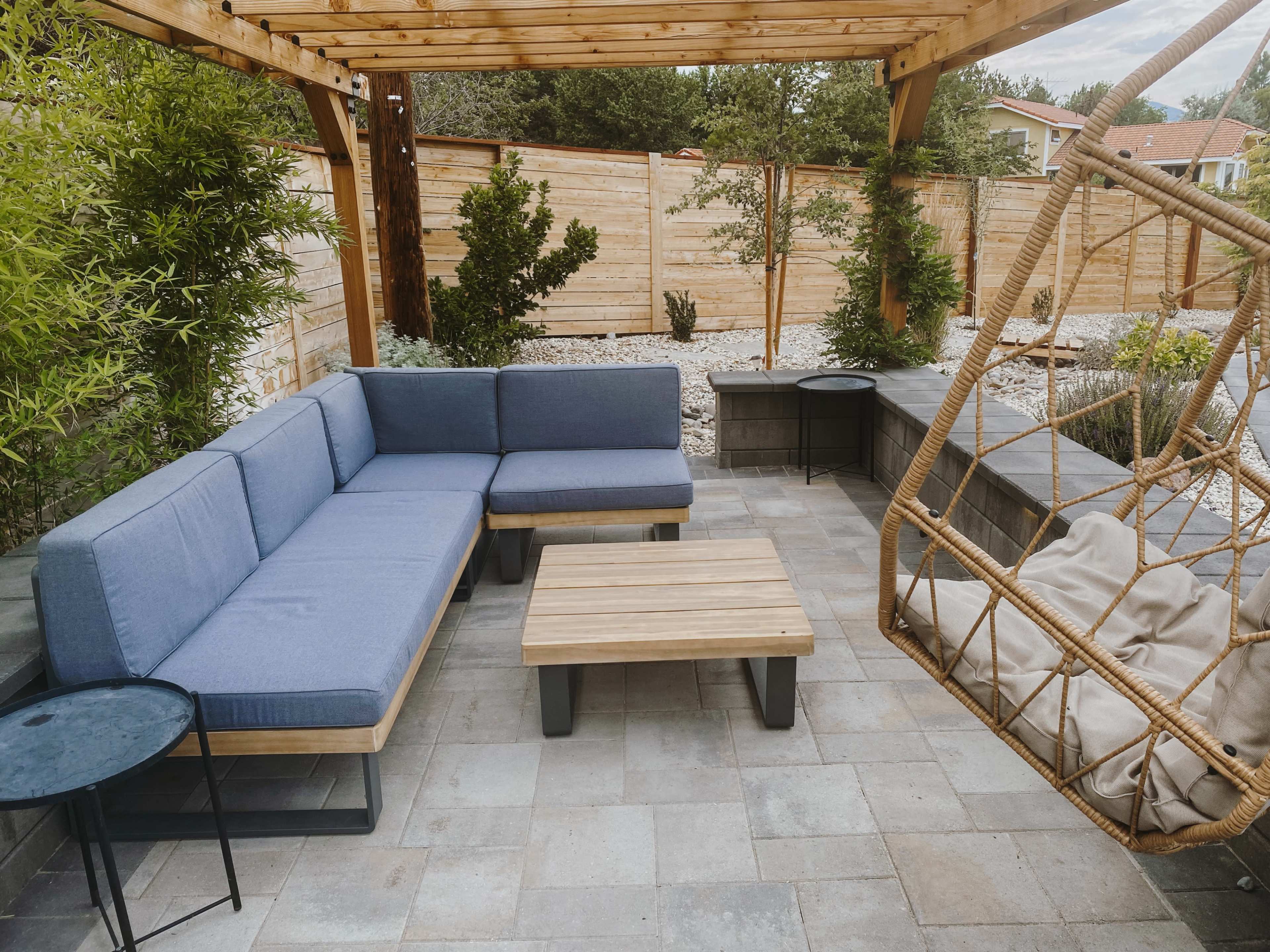 A cozy outdoor seating area featuring a blue sectional couch, a wooden coffee table, and a hanging chair, all surrounded by greenery and stone pathways.