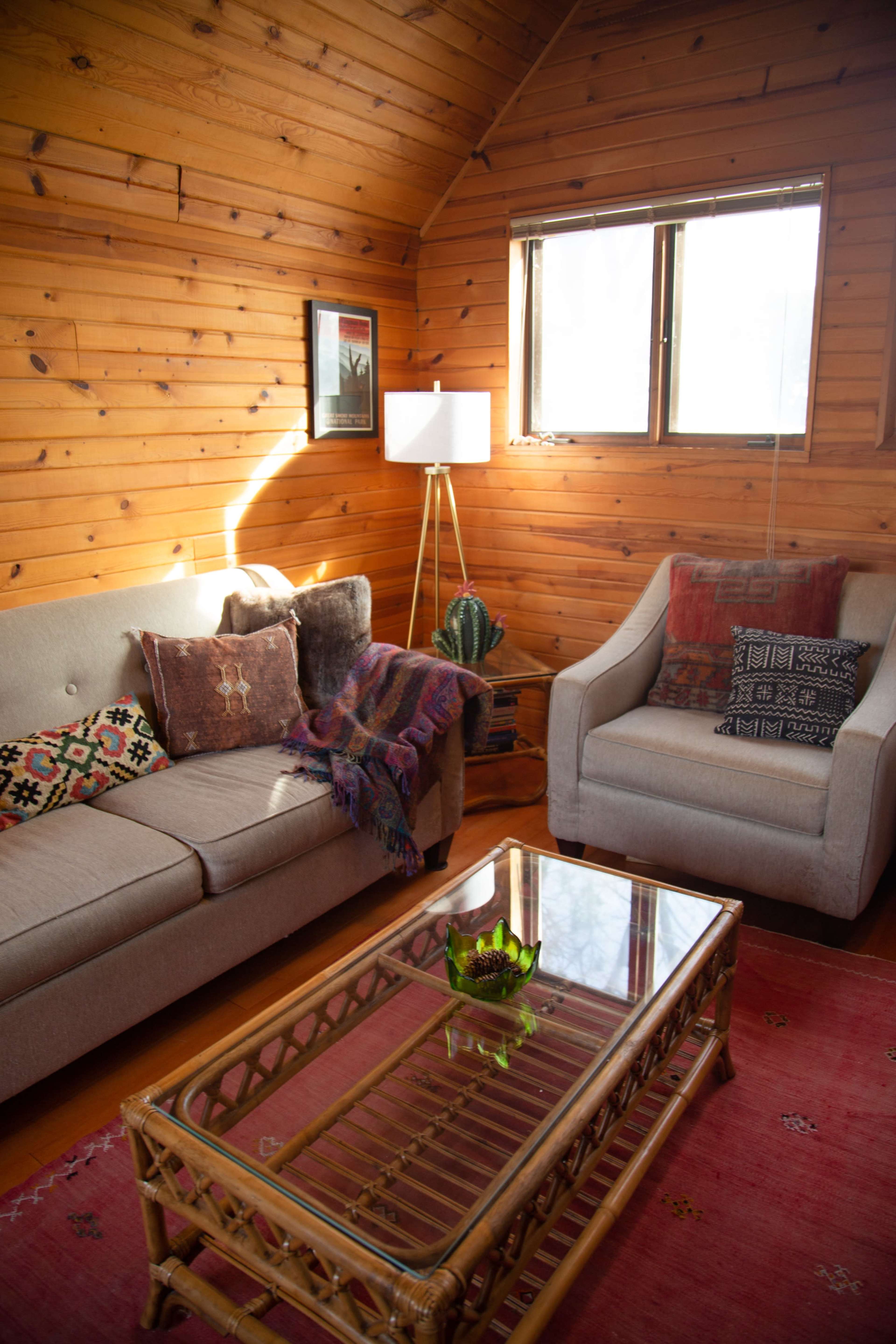 The cozy living room features a beige sofa and a light-colored armchair, surrounded by wooden walls, a glass coffee table, and a floor lamp.