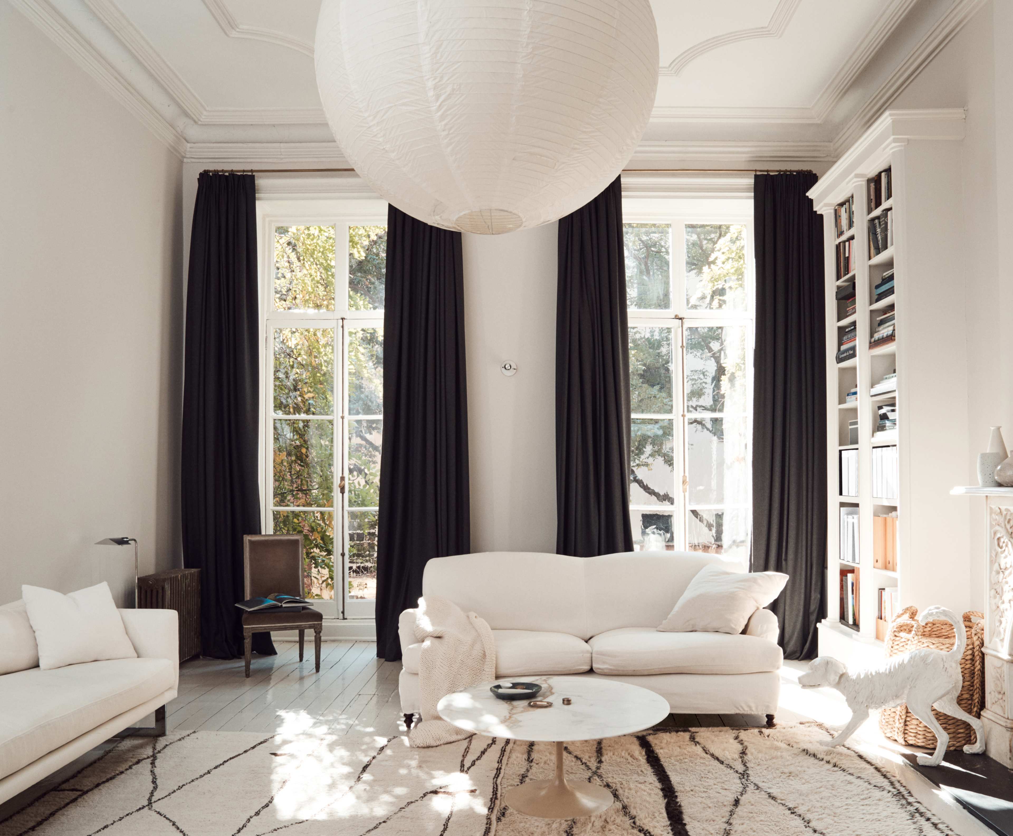 A bright living room with large windows, a white sofa, a marble coffee table, and bookshelves against the wall.