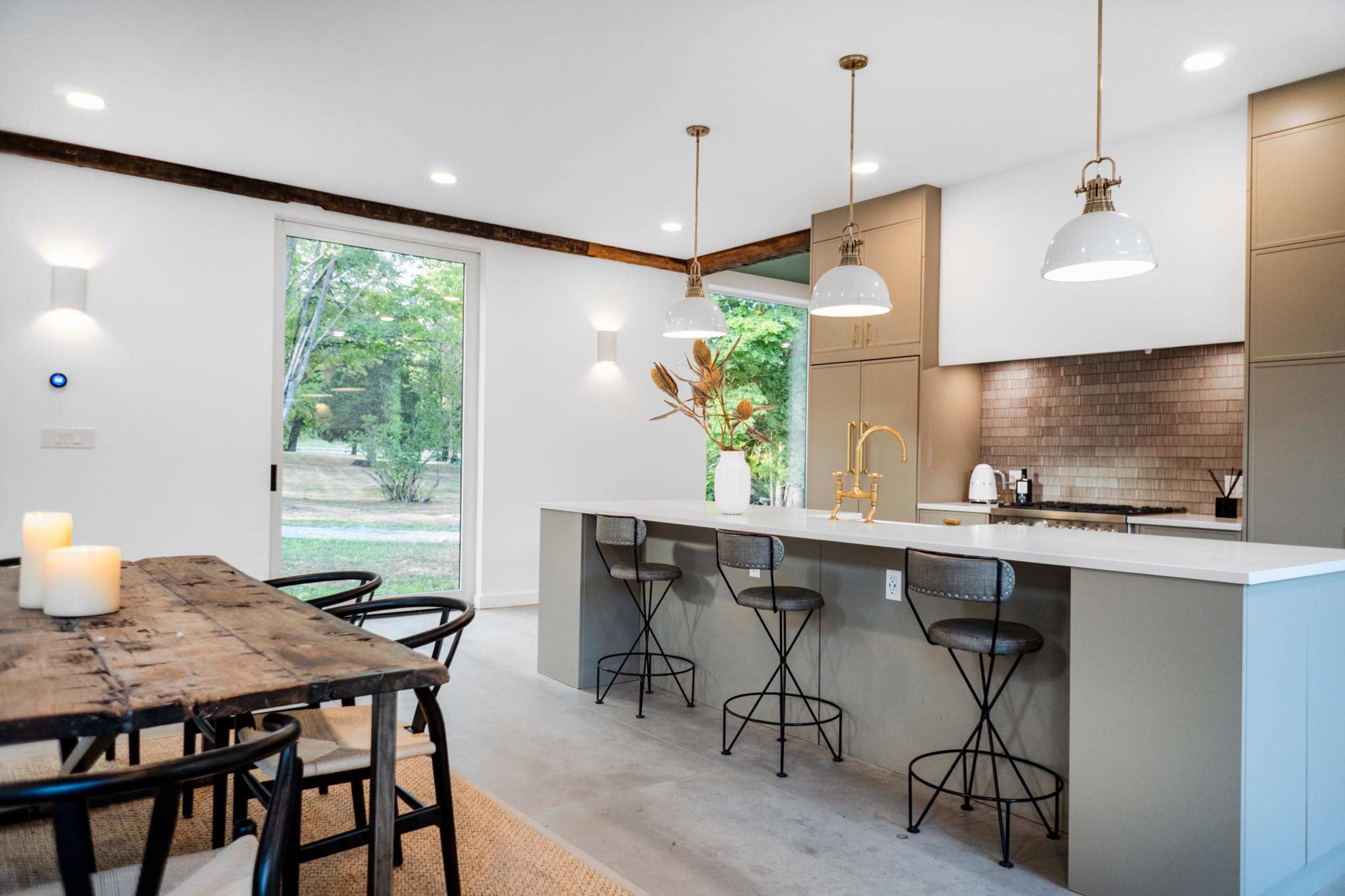 The image features a modern kitchen with a large island, bar stools, and a dining table, surrounded by large windows that allow natural light to illuminate the space.