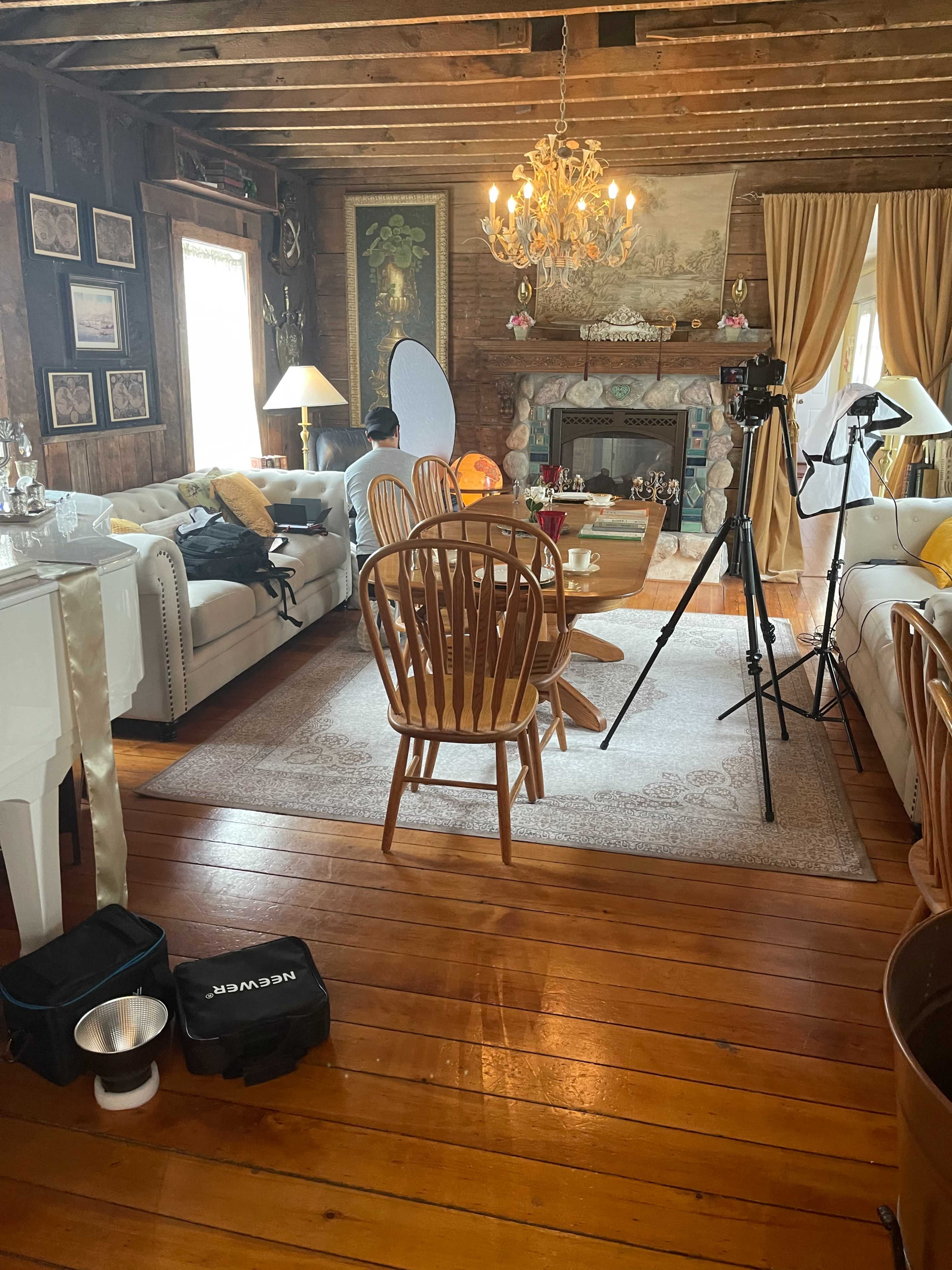 A rustic living room with wooden floors, a chandelier, a fireplace, and a setup for photography, including a camera on a tripod and lighting equipment.