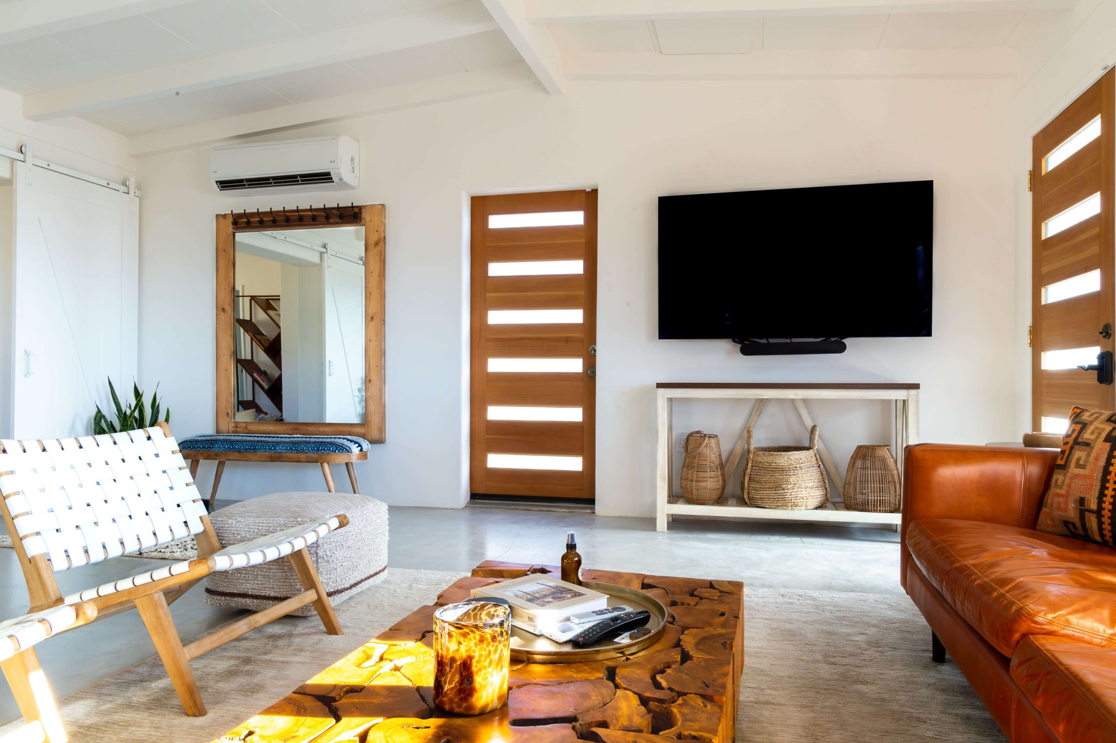 The living room features a leather sofa, a wooden coffee table, and a television mounted on the wall, with a mirror and wooden doors in the background.