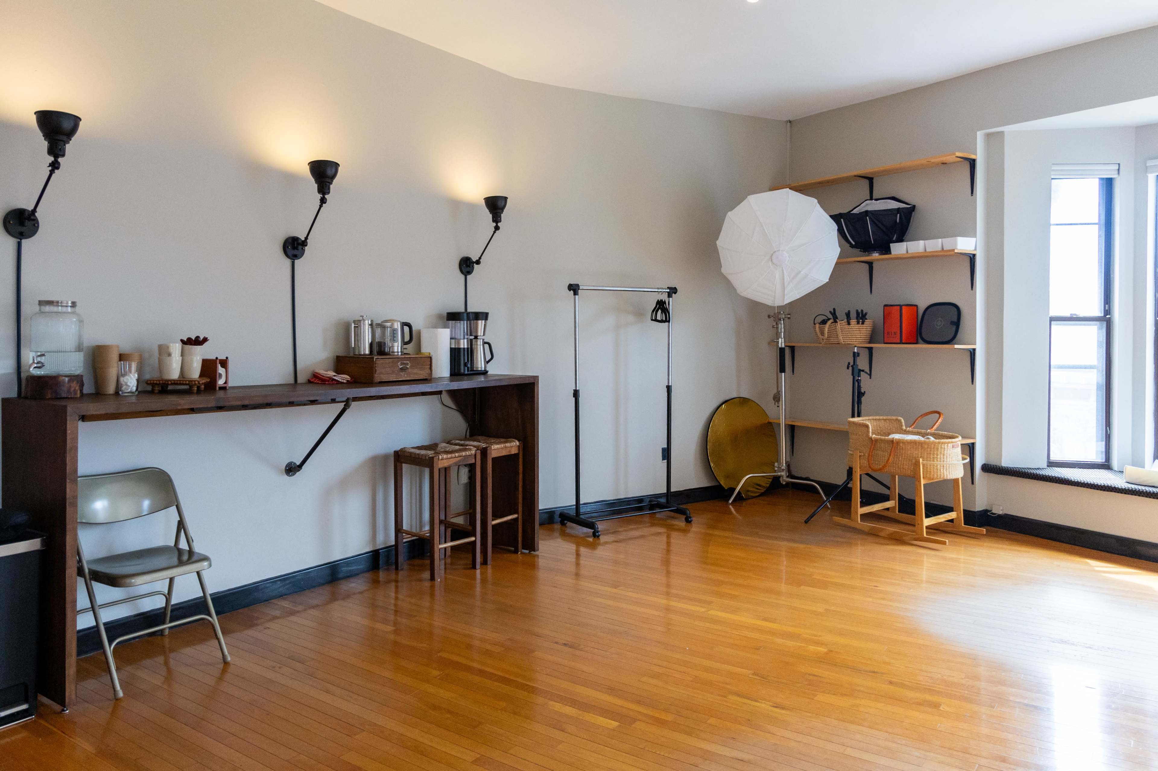 The image shows a minimalist room with hardwood flooring, featuring a wall-mounted shelf, a coffee station, a clothing rack, and a large softbox light set up near a window.