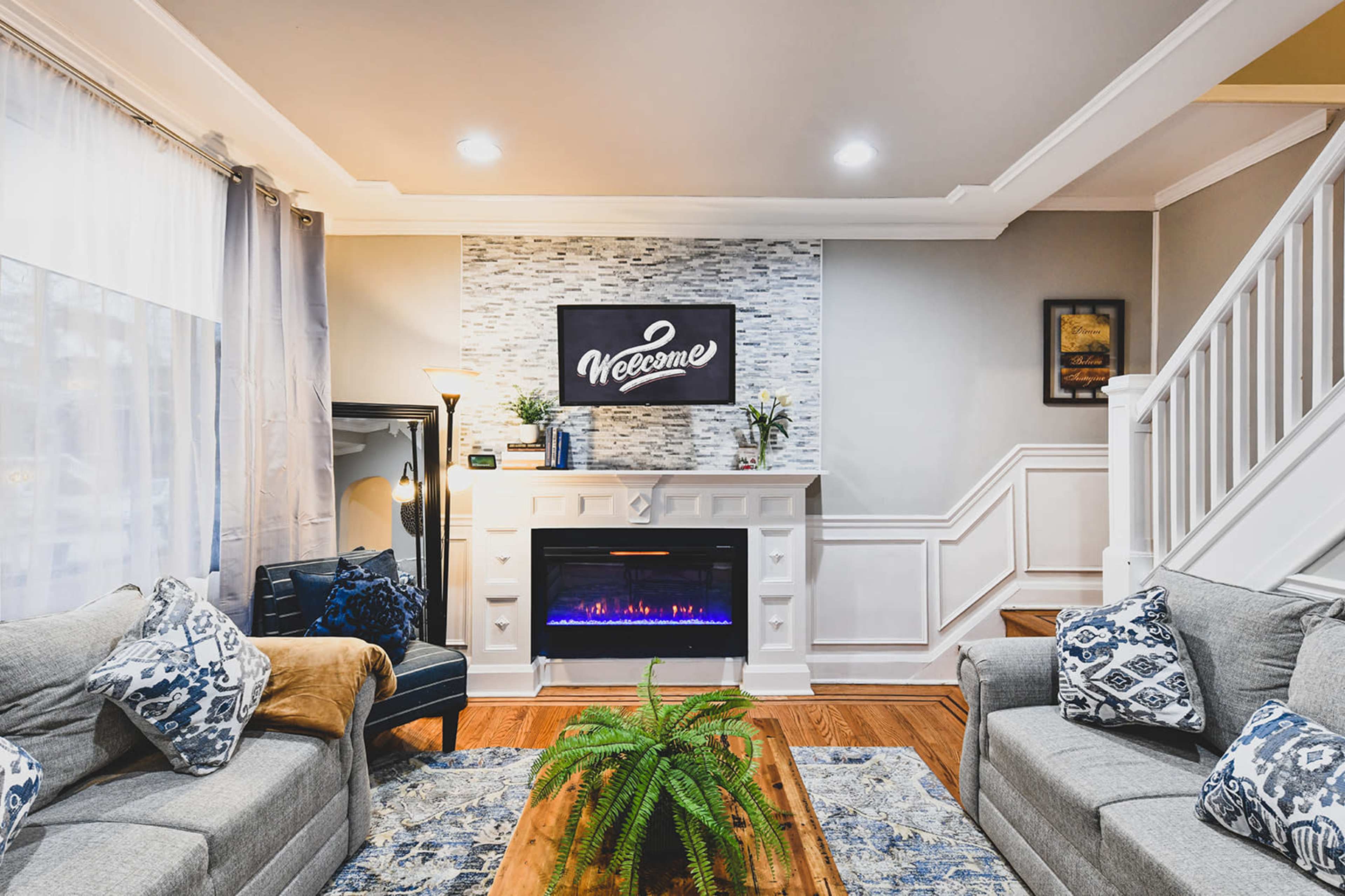 A cozy living room features a television above a modern fireplace, with gray sofas adorned with patterned cushions and a wooden coffee table.