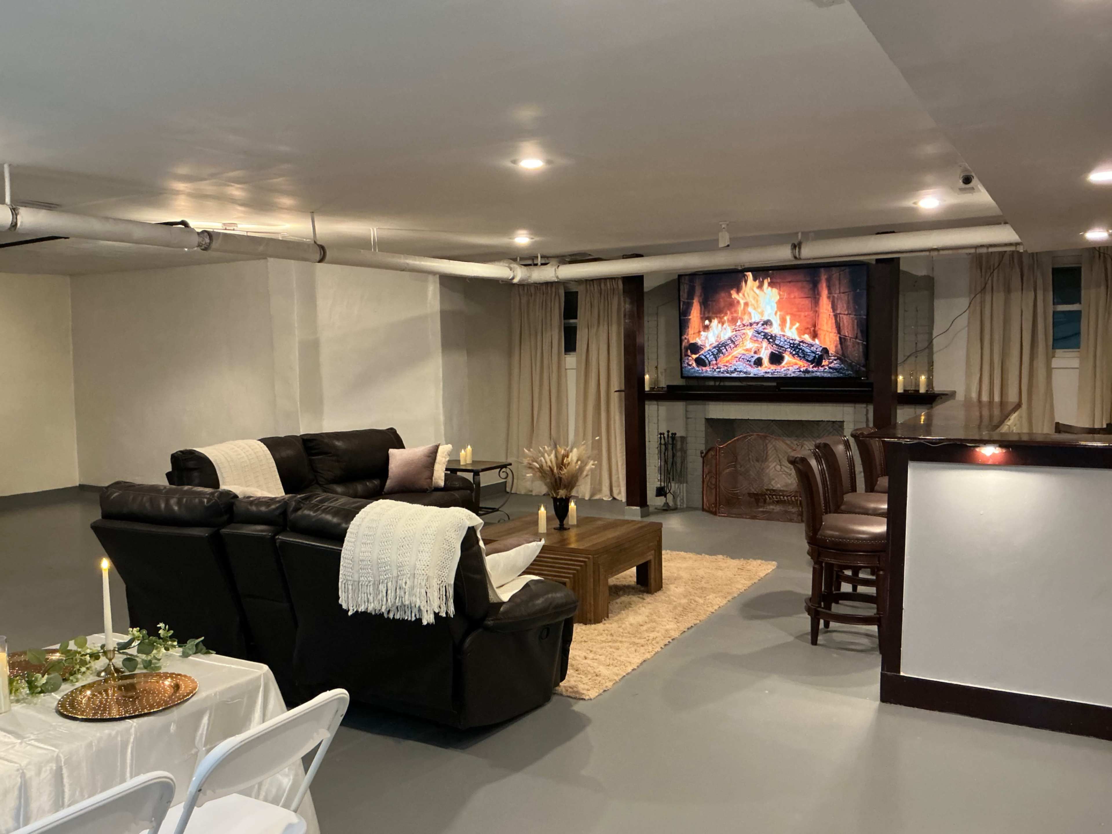 The image shows a basement living area with a leather sectional sofa, a coffee table, a bar with stools, and a large television displaying a fireplace, along with a dining setup.