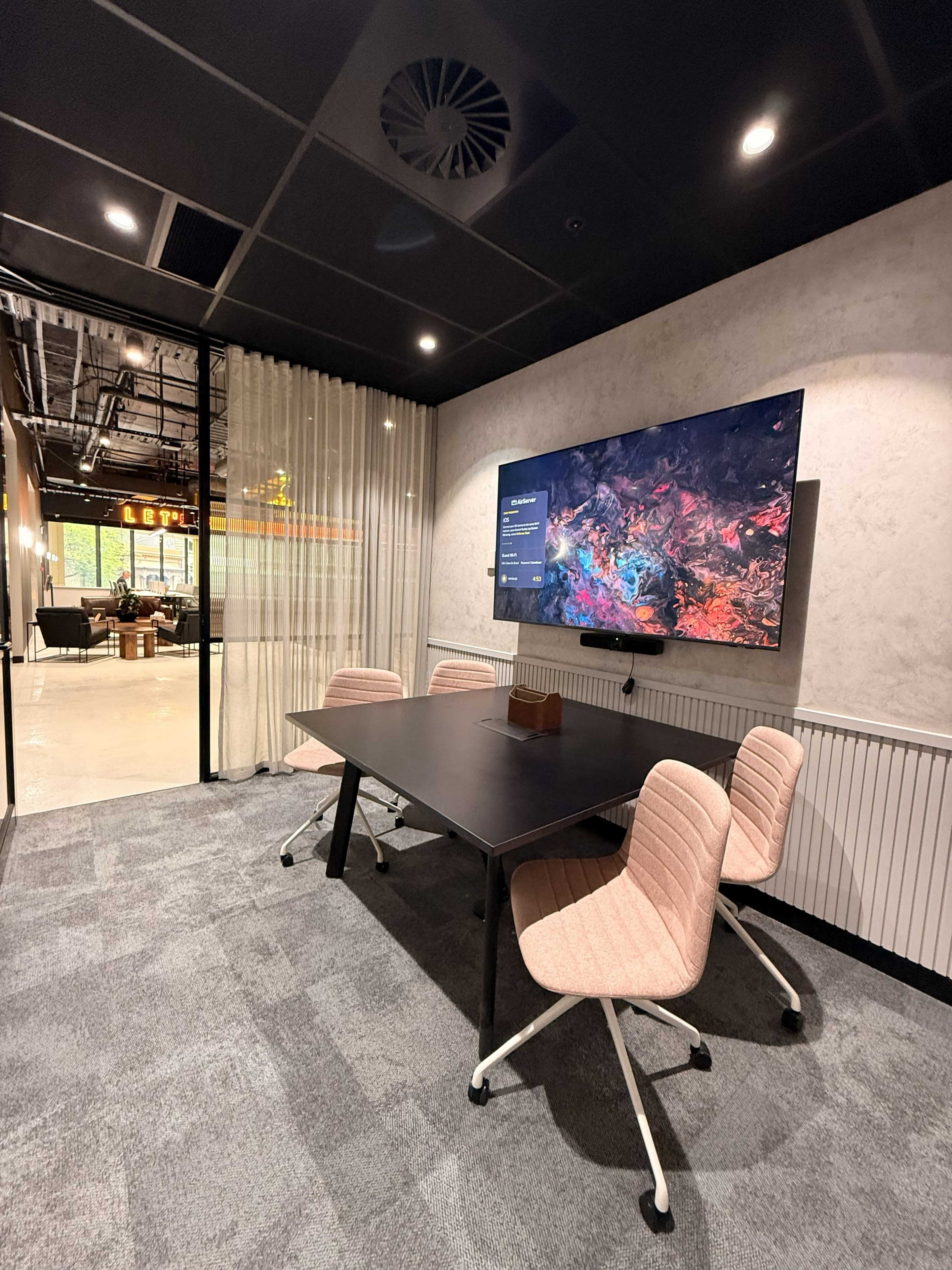 The image shows a modern conference room featuring a table with four pink chairs and a large TV mounted on the wall.