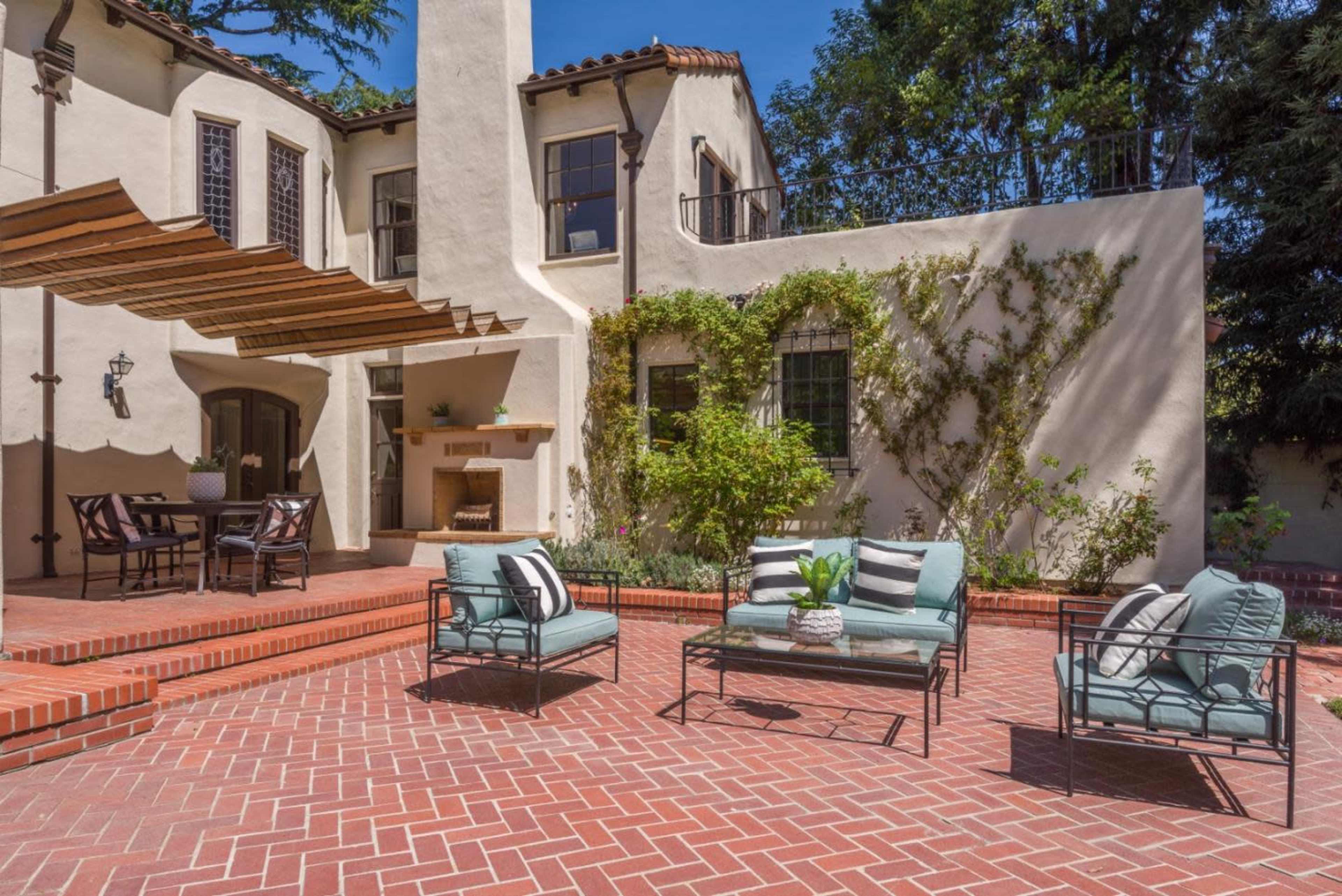 A cozy outdoor patio features comfortable seating and a dining area, surrounded by lush greenery and a charming house.