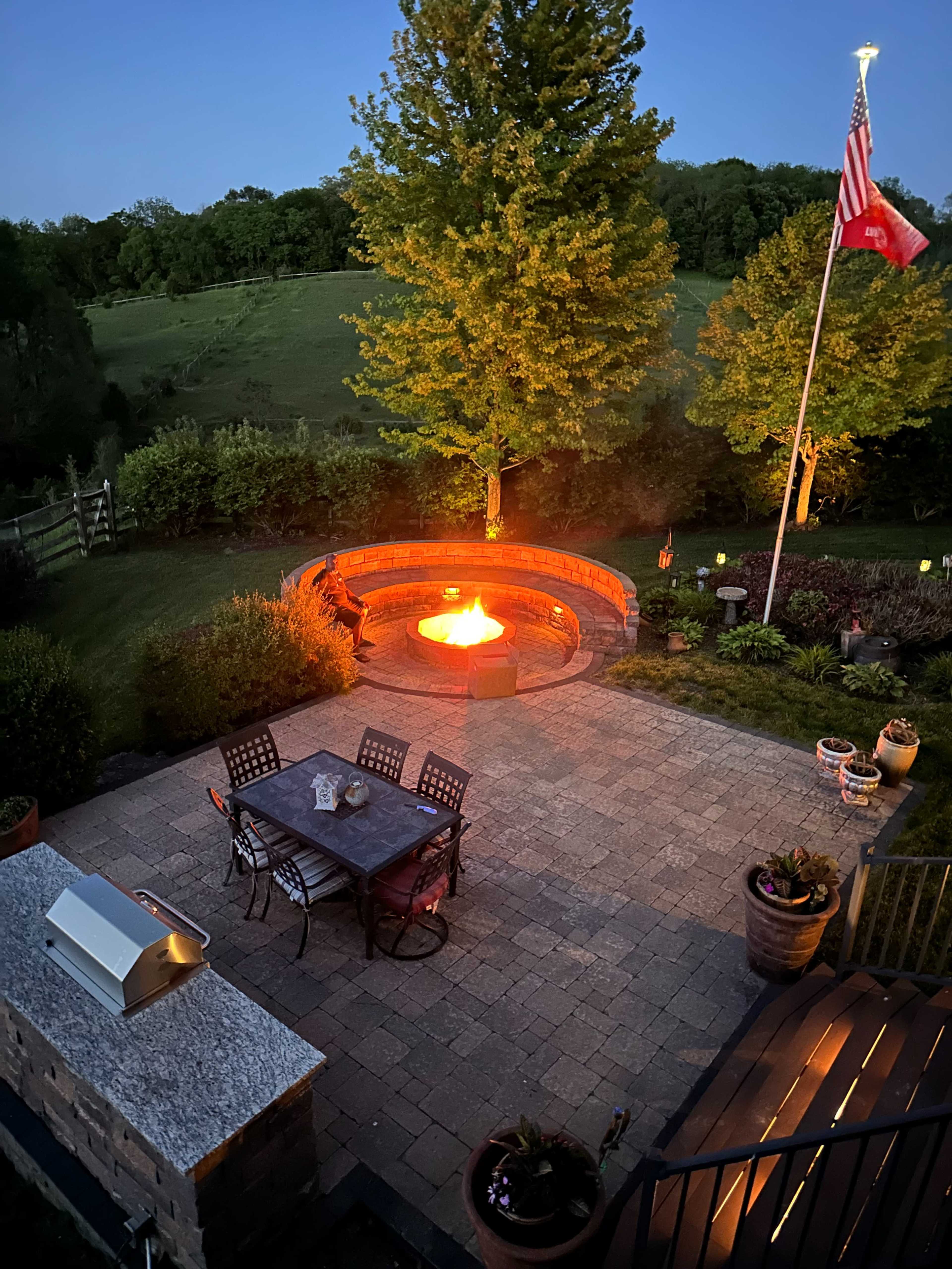 The image shows a stone patio with a fire pit, a table with chairs, and a flagpole displaying an American flag, surrounded by greenery and landscaping at dusk.