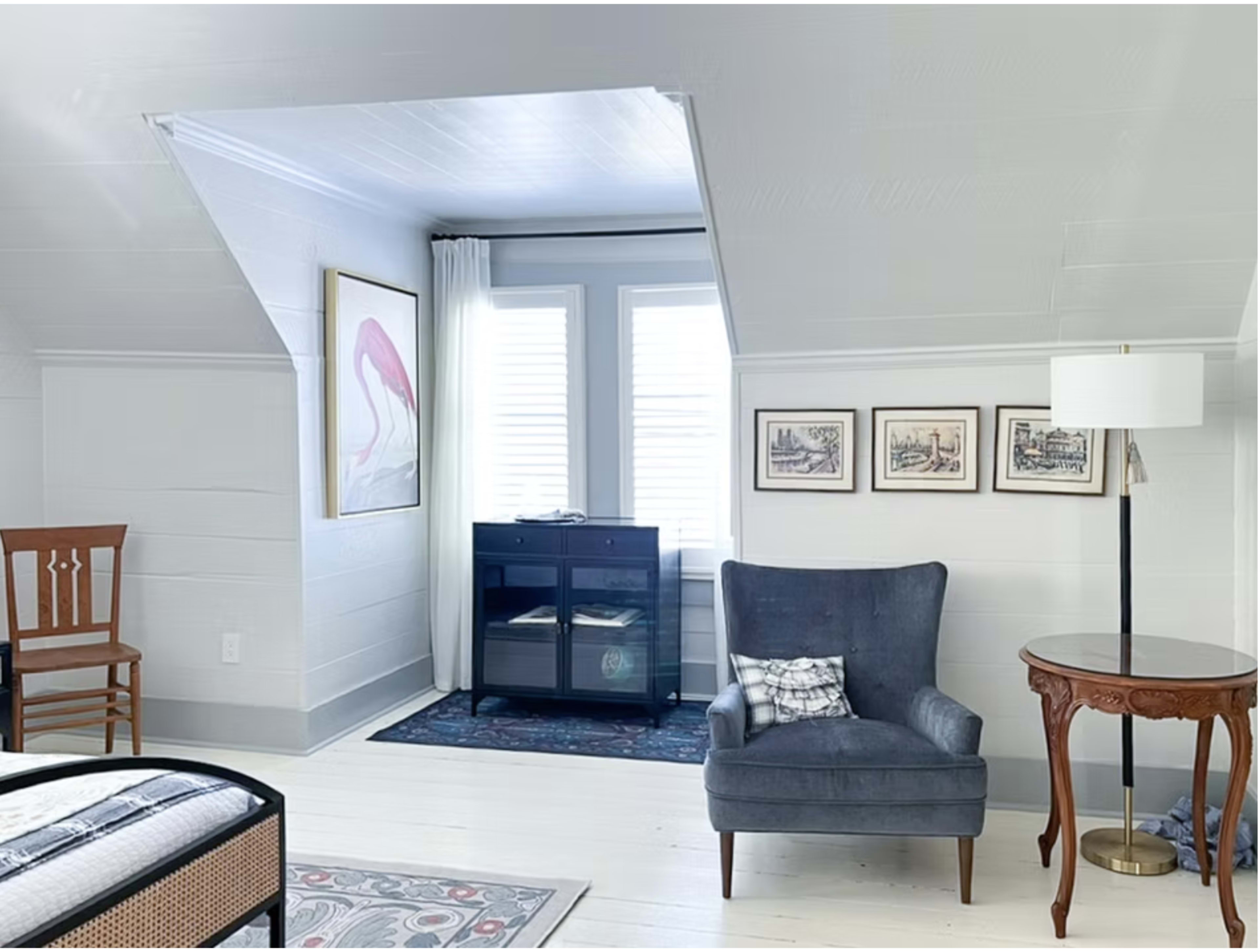 The image shows a cozy attic space featuring a blue armchair, a wooden side table, a lamp, and framed artwork on the walls, with a small black cabinet and a window partially covered by curtains.