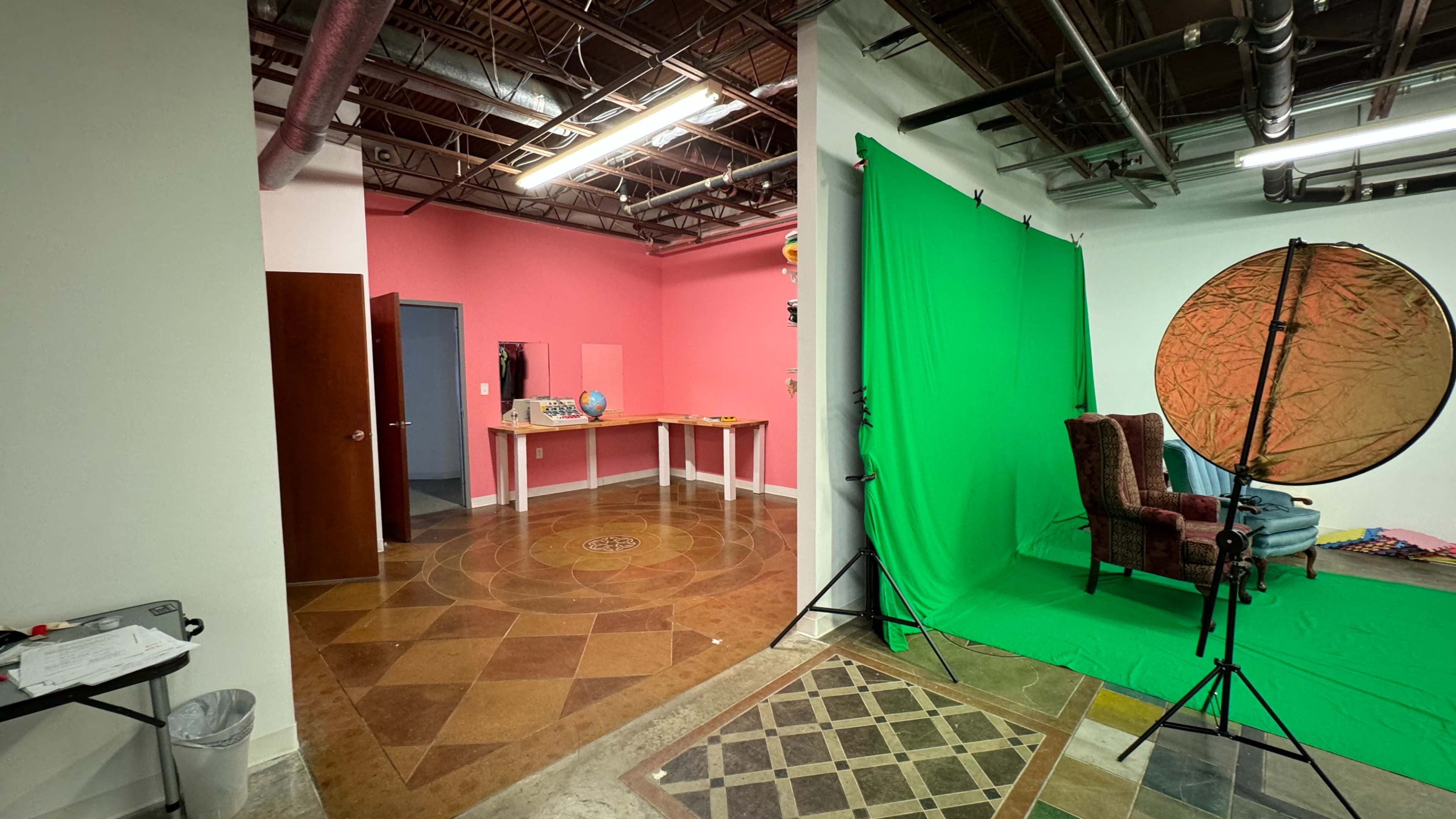 A split scene with a pink-walled room on the left featuring a table and a globe, and a green backdrop setup on the right with a patterned floor.