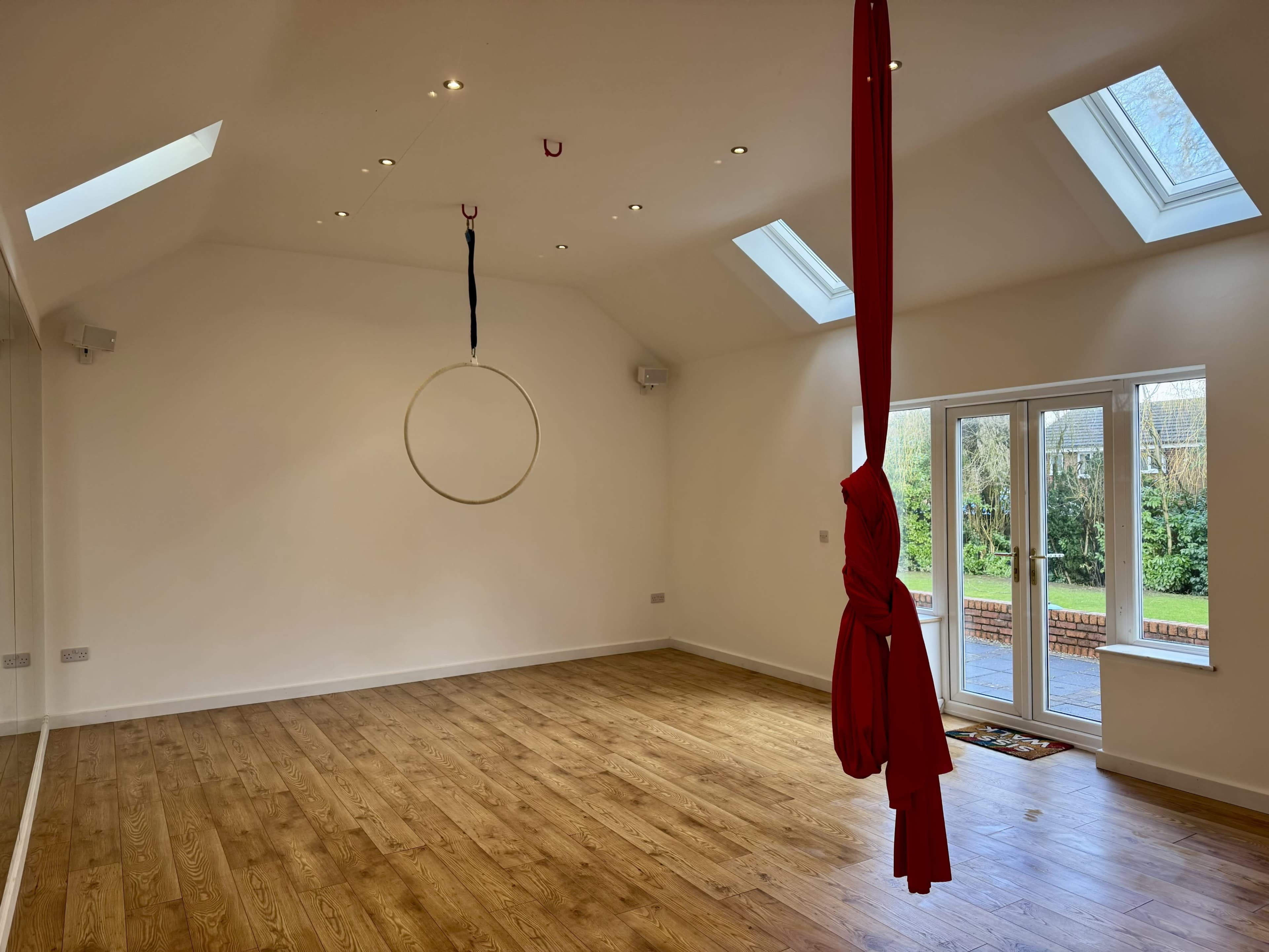 The image shows a spacious room with wooden flooring, two skylights, a circular aerial hoop hanging from the ceiling, and a red draped fabric.