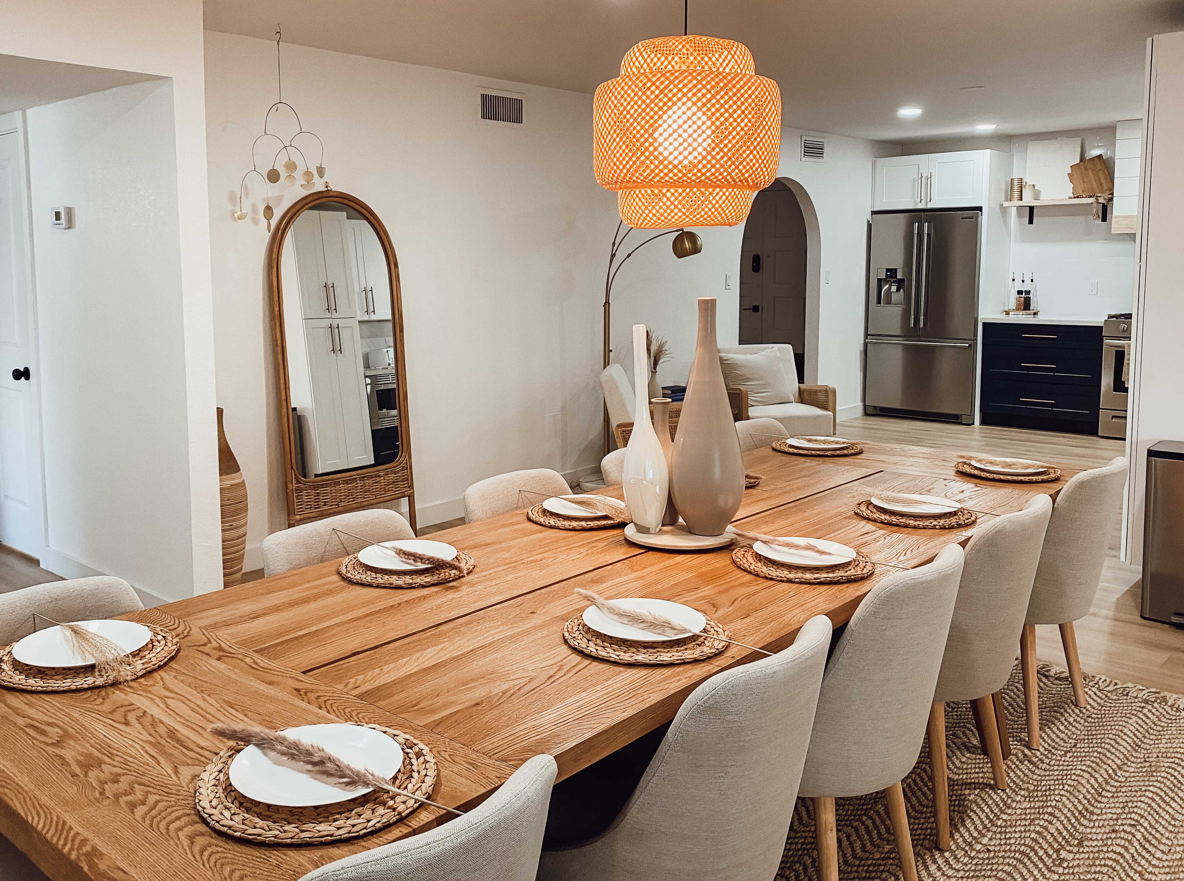 A dining room features a long wooden table set with plates and woven placemats, illuminated by a large woven pendant light, with a mirror and a modern kitchen visible in the background.