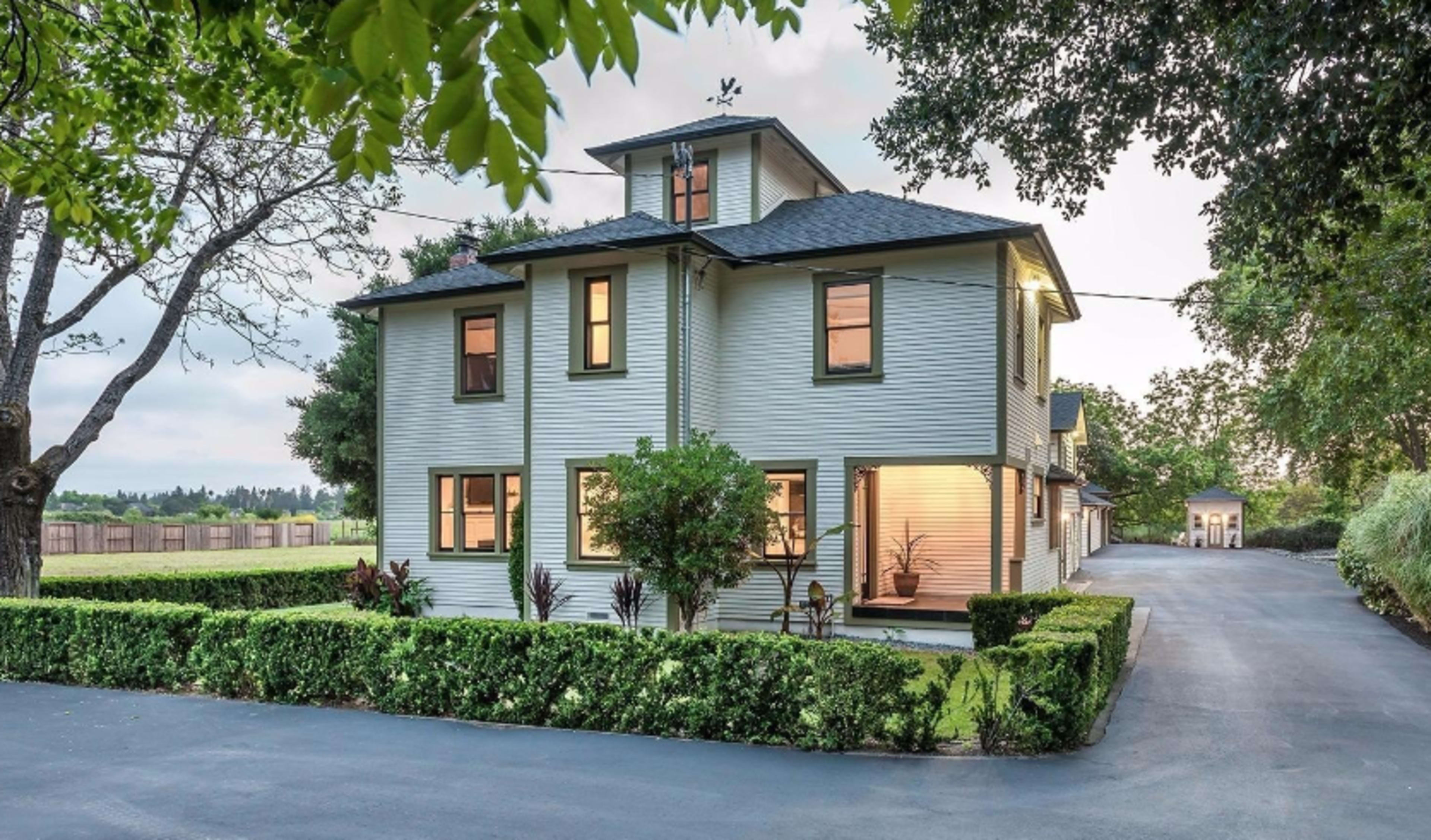 A two-story house with a distinctive design stands surrounded by well-manicured hedges and a paved driveway.