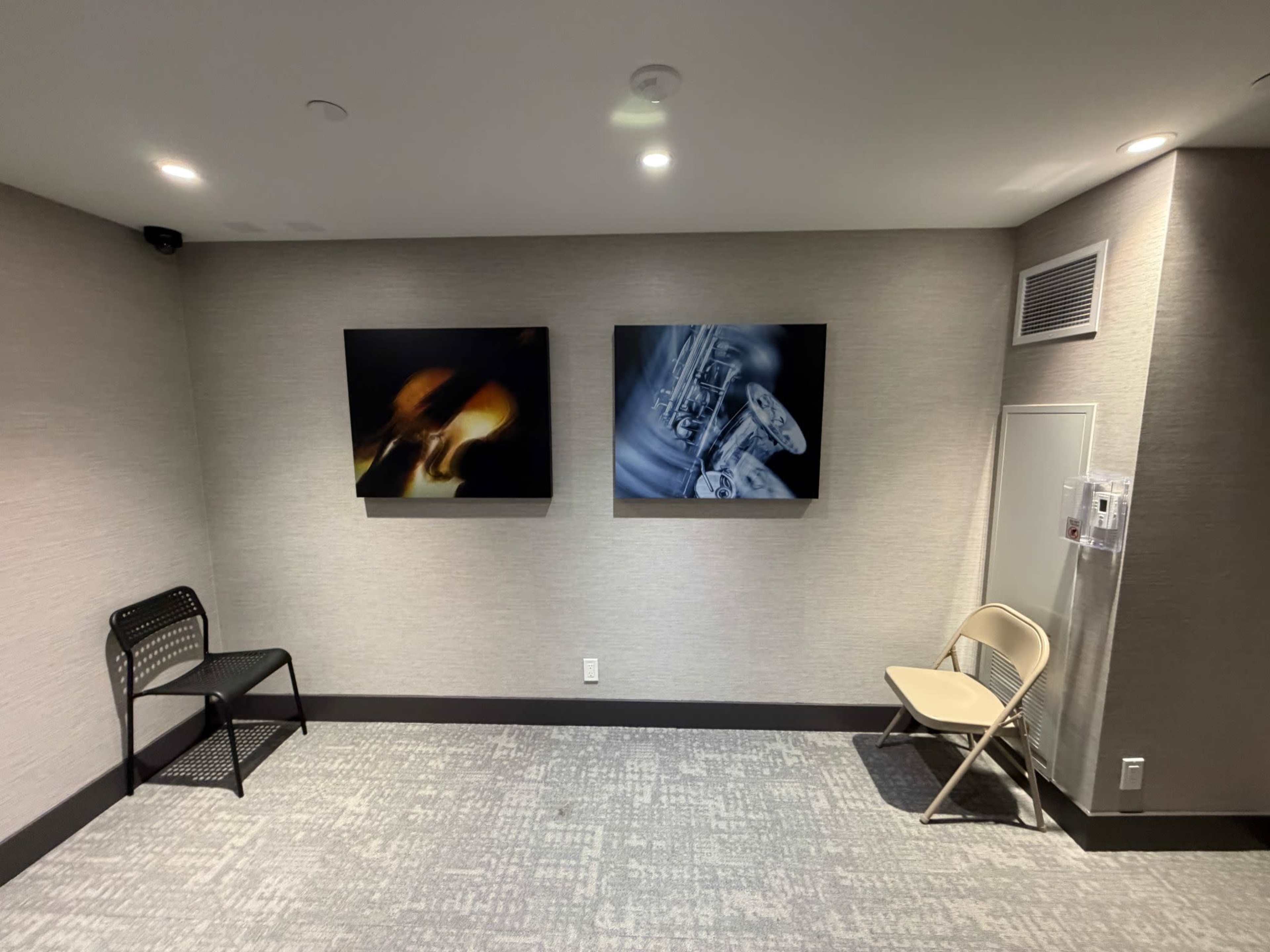 The image shows a corner of a room with two abstract paintings on the wall, along with a black chair and a folding chair on the floor.