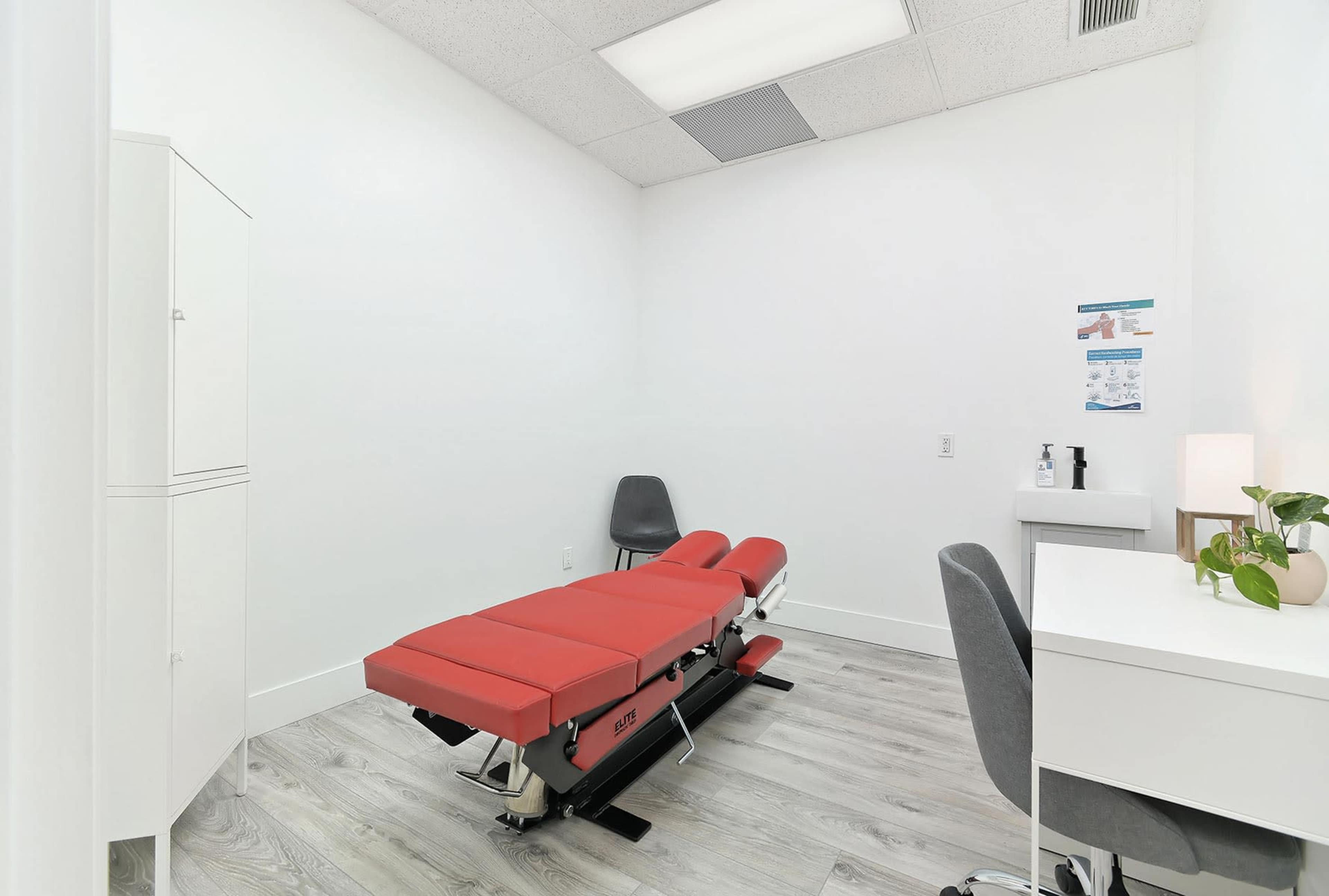 A clean therapy room featuring a red treatment table, a desk with a plant, and a gray chair.