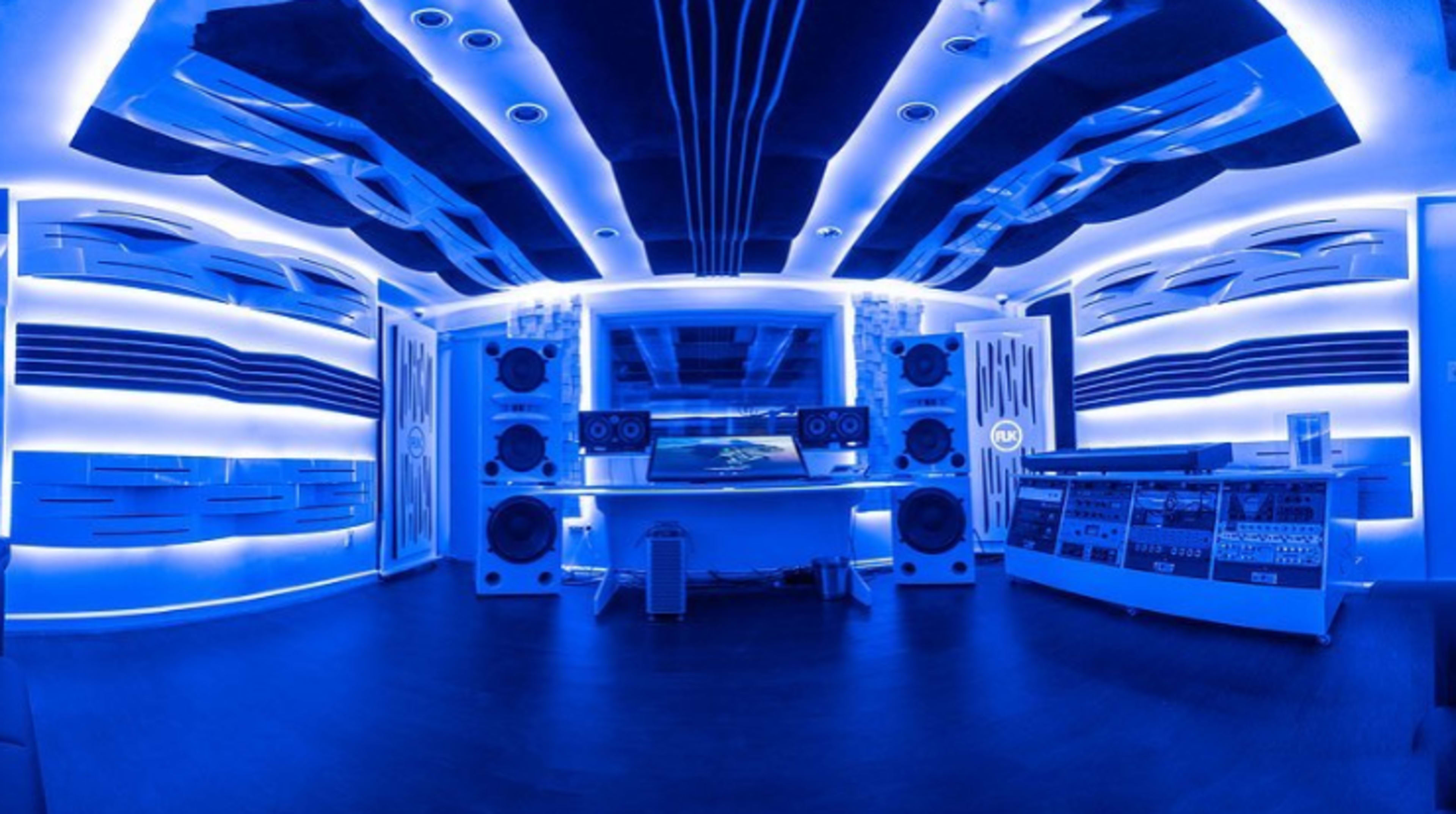 A modern music studio with blue ambient lighting, featuring large speakers and soundproofing panels on the walls.