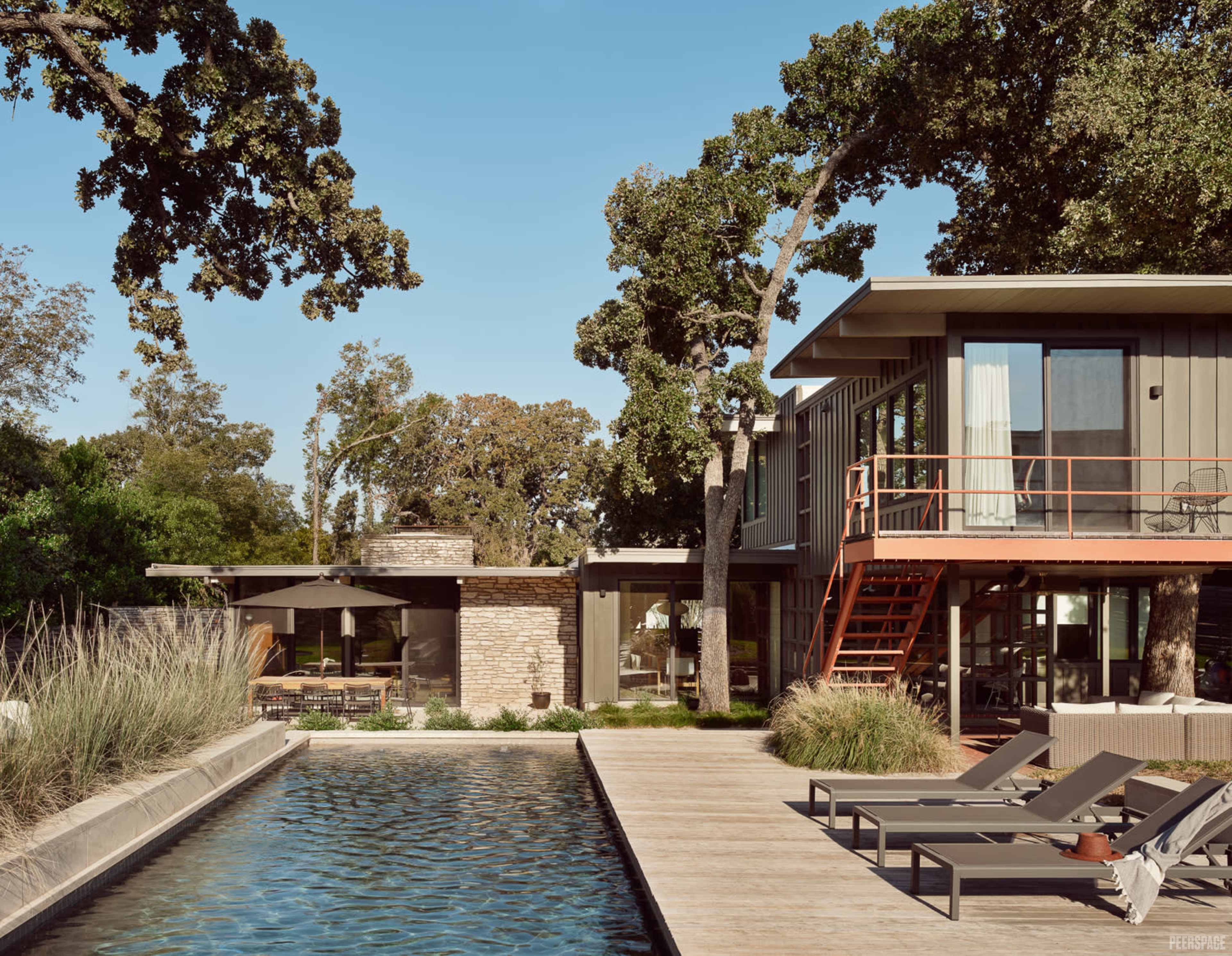 A modern house with large glass windows is situated beside a pool and surrounded by trees and landscaped areas.