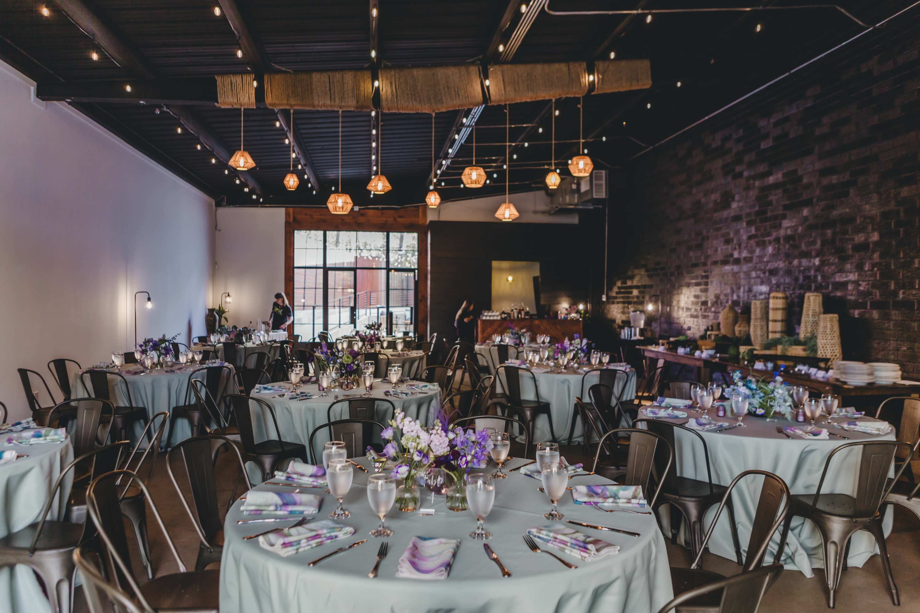 The image shows a beautifully arranged dining area with round tables set for an event, featuring floral centerpieces and decorative tableware under soft lighting.