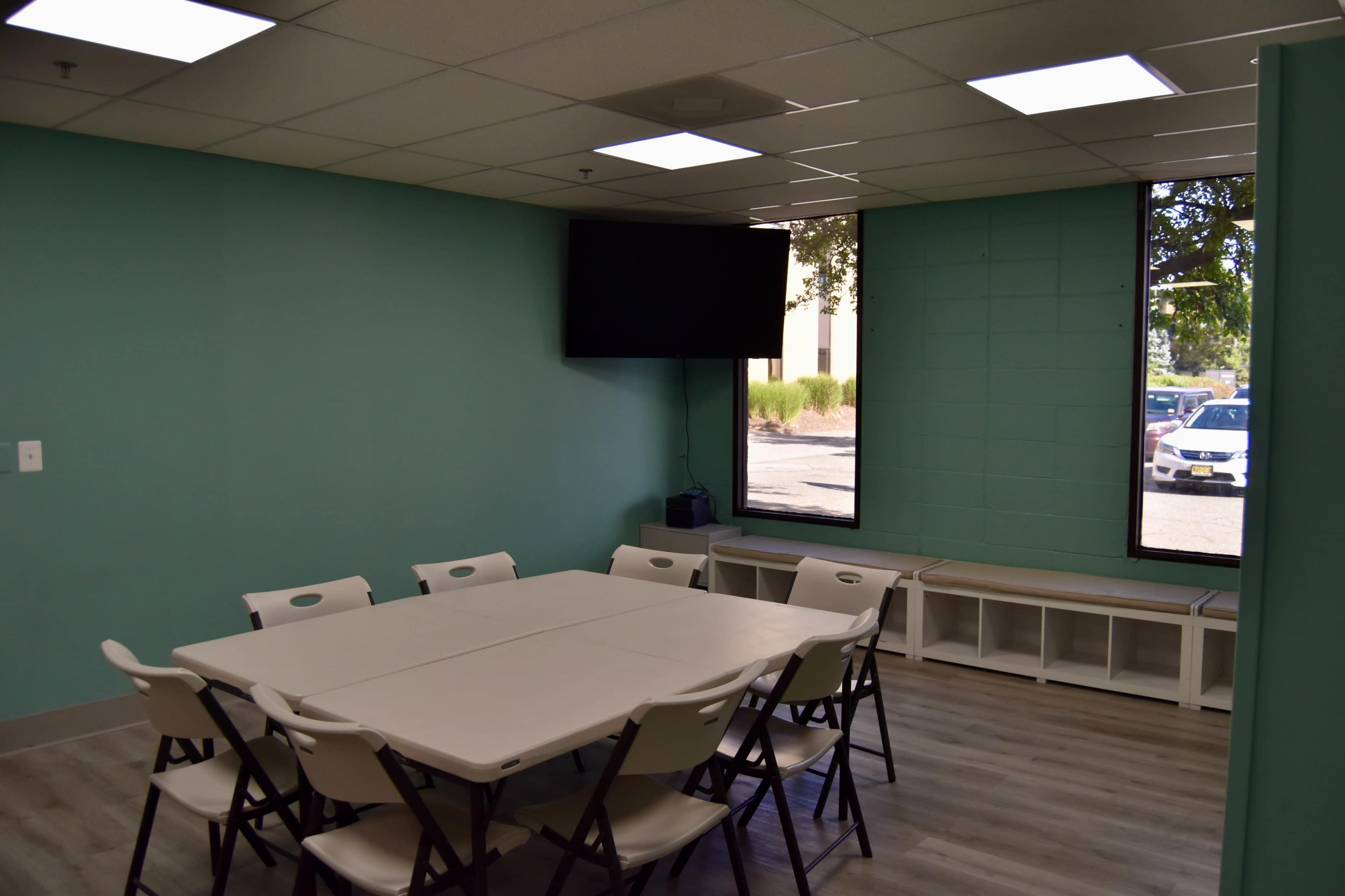 A meeting room features a rectangular table surrounded by eight folding chairs, with a mounted television on the wall and large windows allowing natural light.