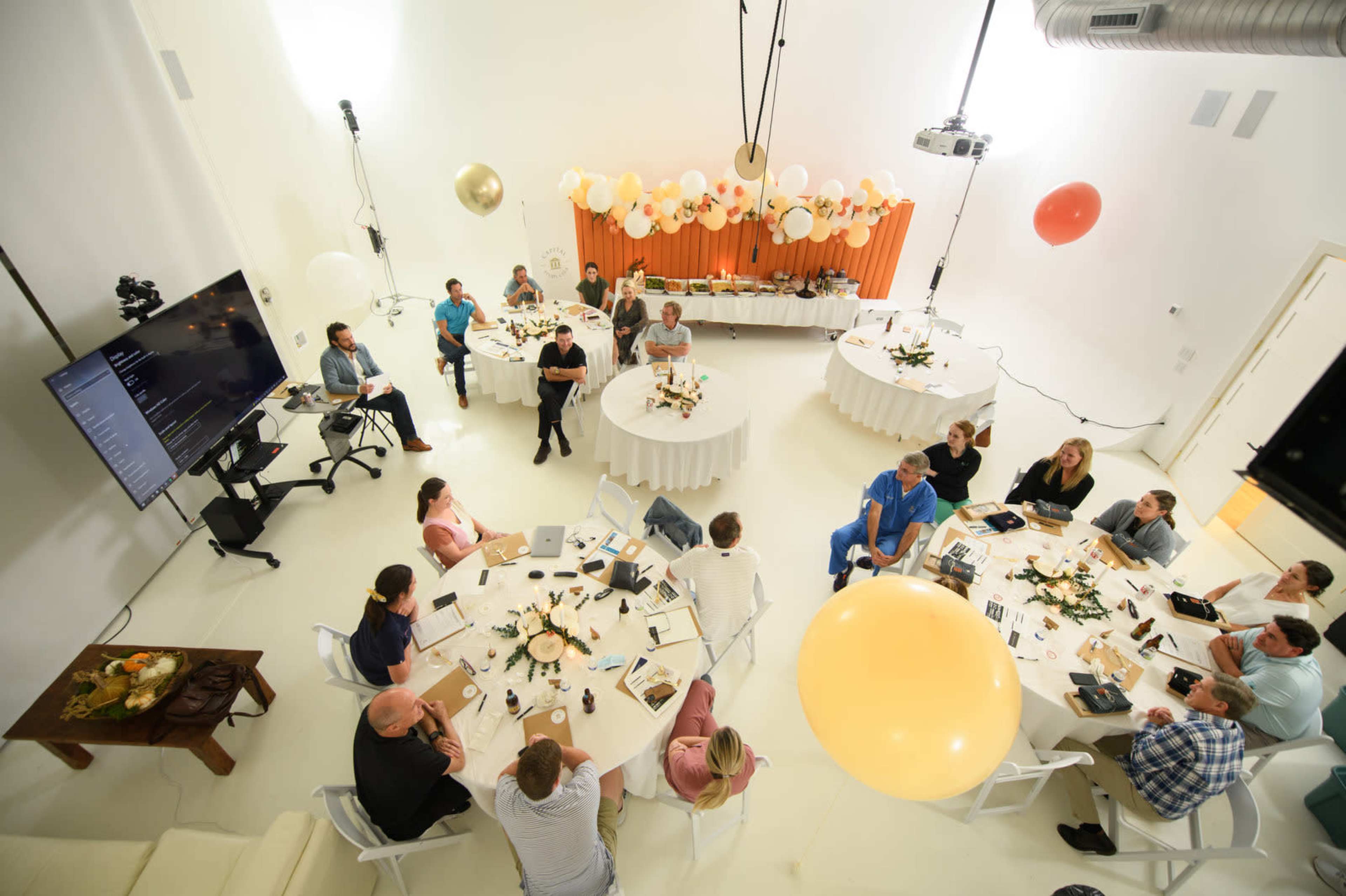 A round table meeting is taking place in a bright, minimalistic room with decorations including balloons and floral arrangements.