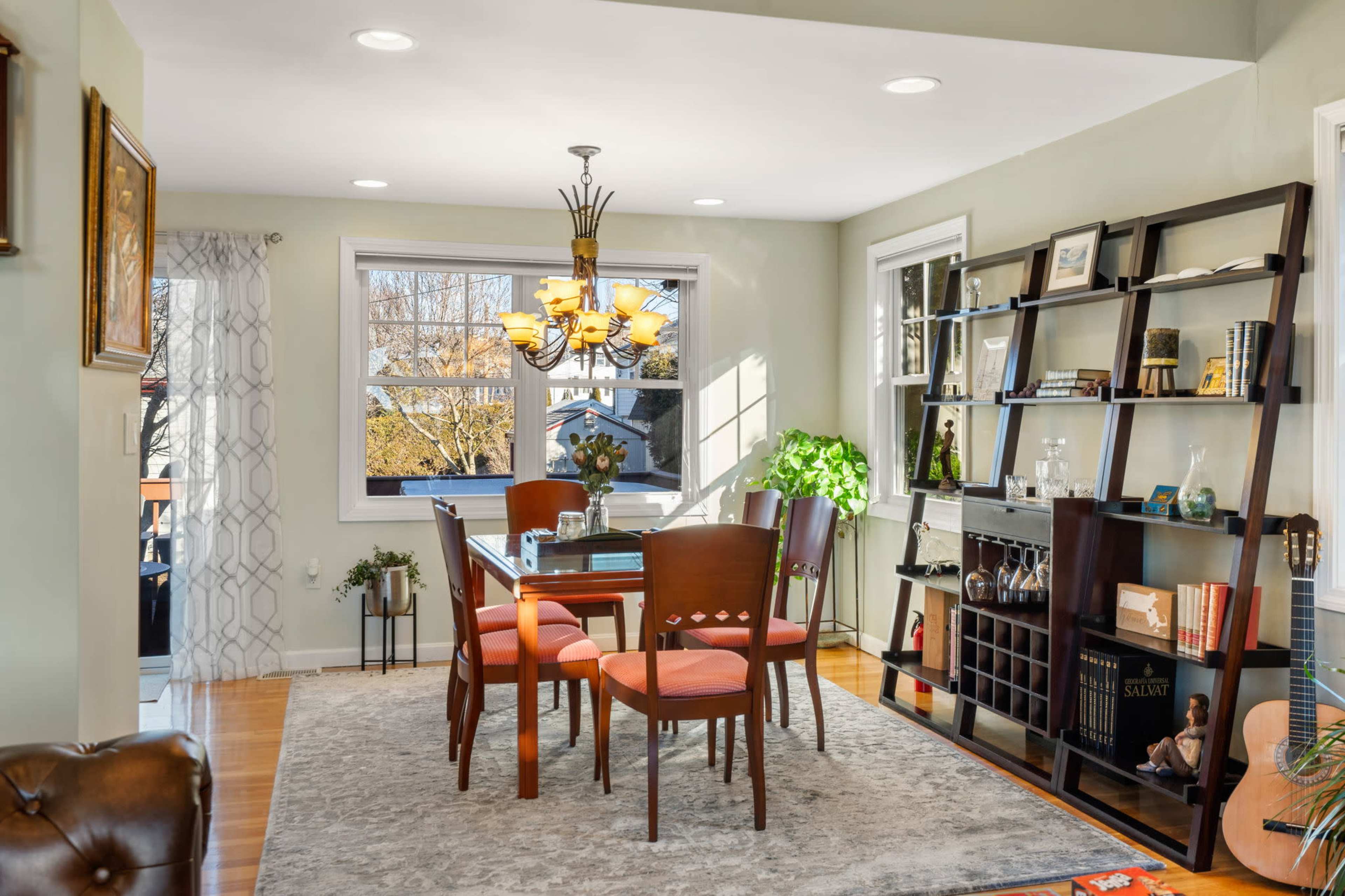 A well-lit dining area featuring a wooden table surrounded by four chairs, a bookshelf filled with various items, and large windows allowing natural light to enter.