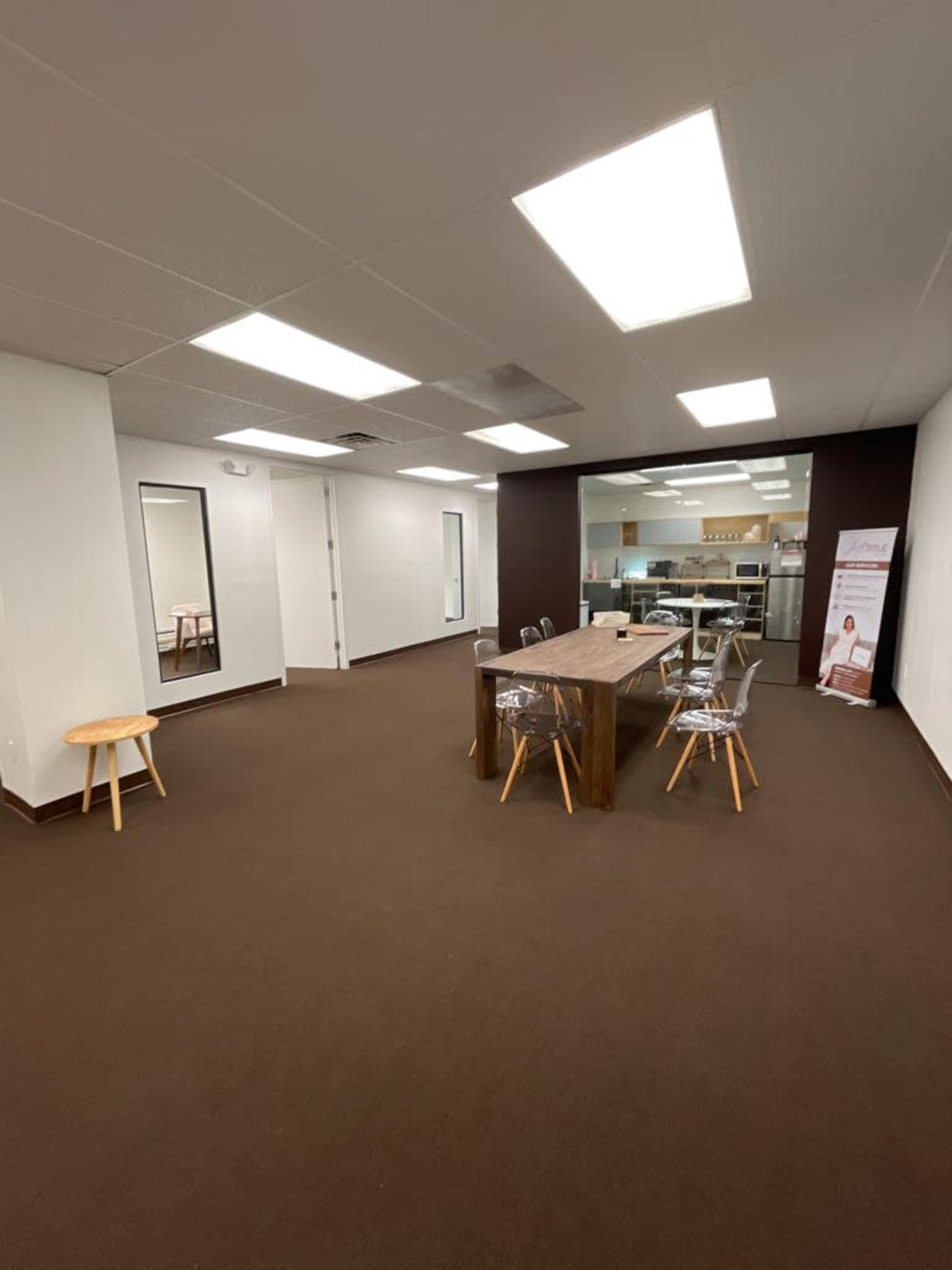 A minimalist office space featuring a large wooden table surrounded by clear chairs, with a mirror and a kitchenette visible in the background.