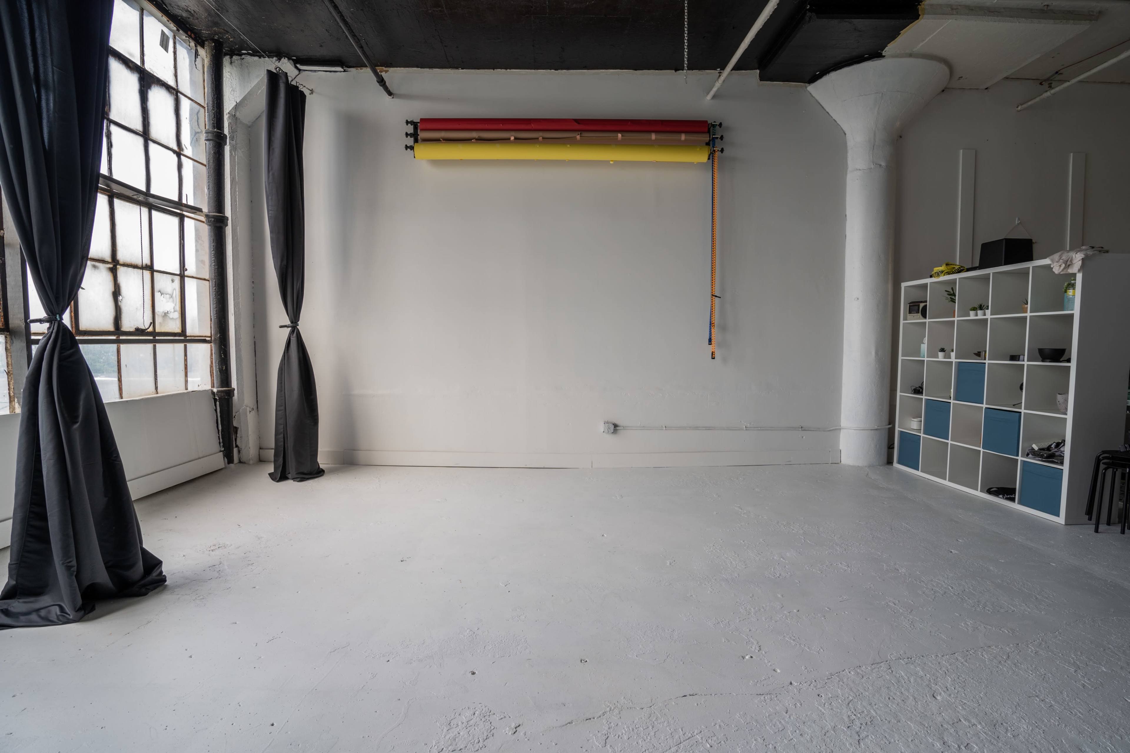 A spacious, minimalist studio with gray walls, a large window draped with black curtains, and a white shelving unit against one wall.